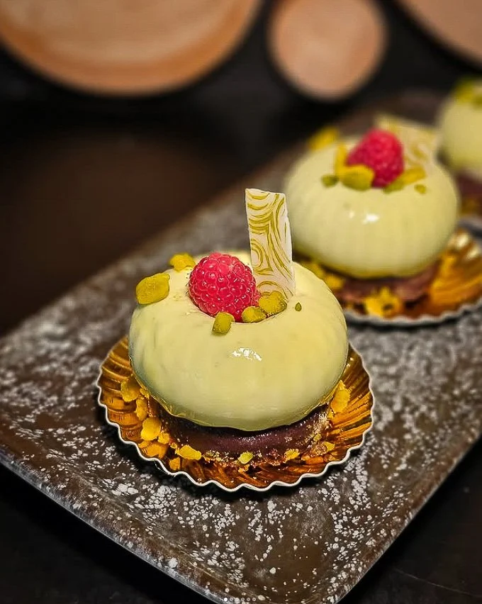 Mandelguss-Törtchen mit weißer Glasur, roter Beere, Pistazien und weißer Schokolade auf einem dunklen Teller