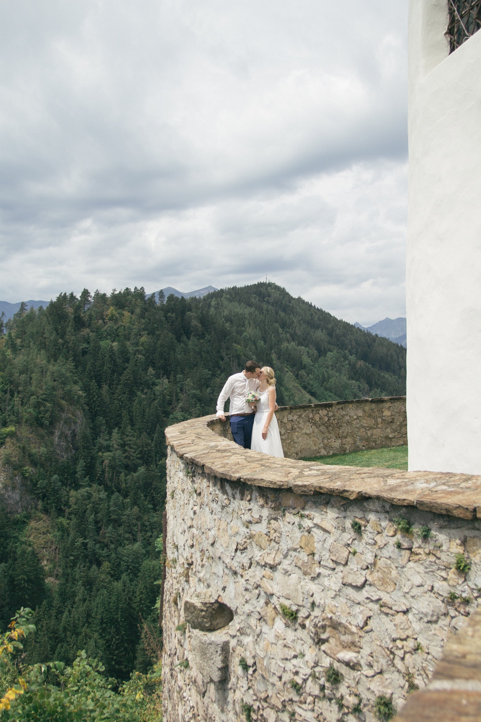 Ein Paar, das auf einer steinernen Balkonmauer steht, vor einer Berglandschaft mit bewaldeten Hügeln und einer bewölkten Himmel stattfindet. Es scheint ein romantisches oder symbolisches Ereignis wie eine Hochzeit zu sein.