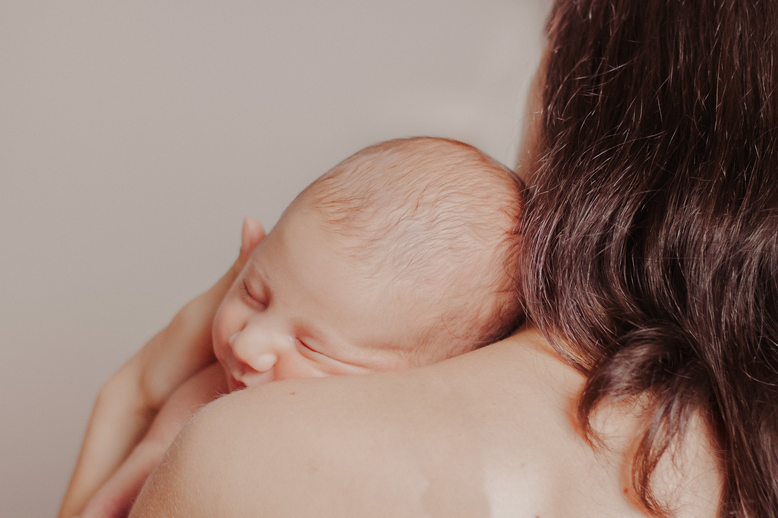 Nahaufnahme einer schlafenden Baby-Haut, die in den Armen einer Frau liegt, mit braunen Haaren im Hintergrund.