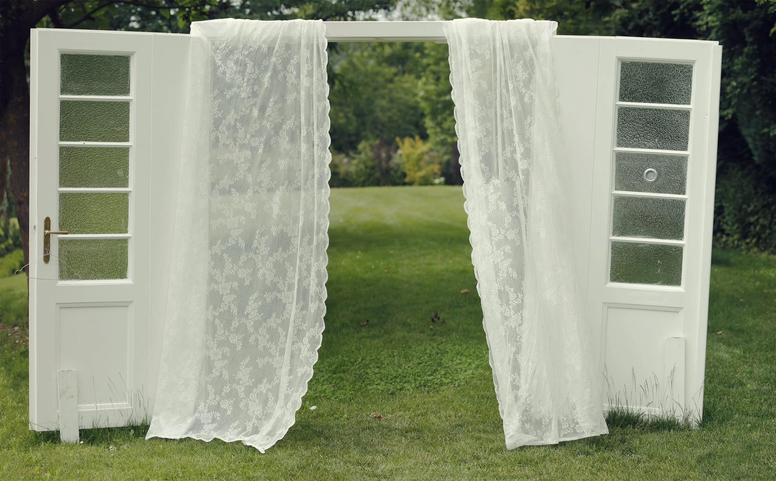 Weiße Fenster- und Türrahmen mit durchsichtigen Vorhängen, Blick auf einen grünen Garten mit Bäumen und Rasen im Hintergrund.