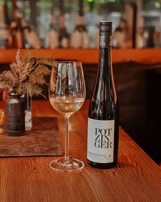 Ein Glas Weißwein und eine Flasche Wein auf einem Holztisch in einem rustikalen Restaurant, im Hintergrund Dekorationen aus Zweigen und Flaschen, warmes Licht.