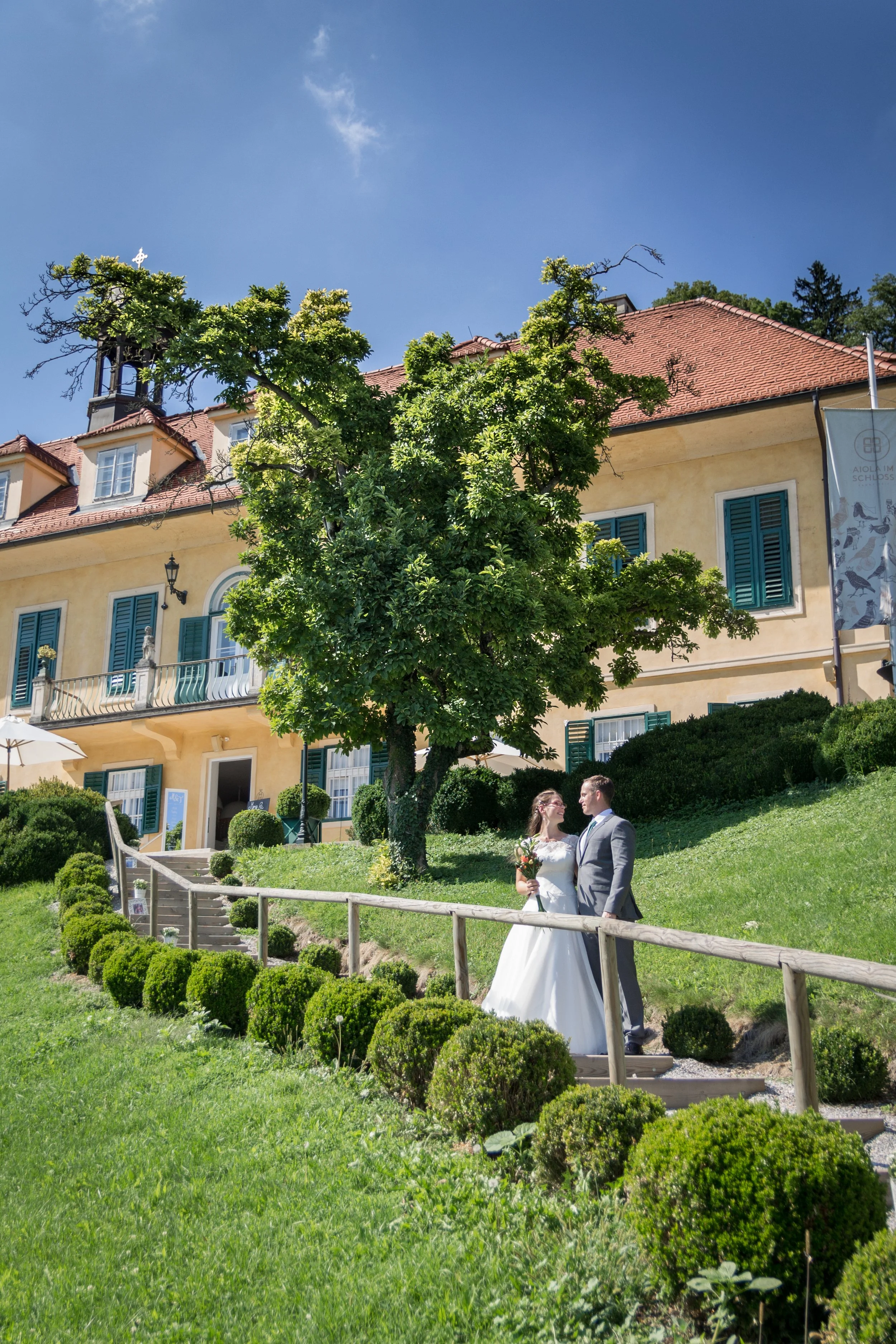 Braut und Bräutigam stehen auf einer Wiese vor einem historischen Gebäude mit gelber Fassade, grünen Fenstern und einem roten Ziegeldach, während sie sich ansehen. Ein Baum und kleine Sträucher sind im Vordergrund sichtbar, im Hintergrund ein blauer 
