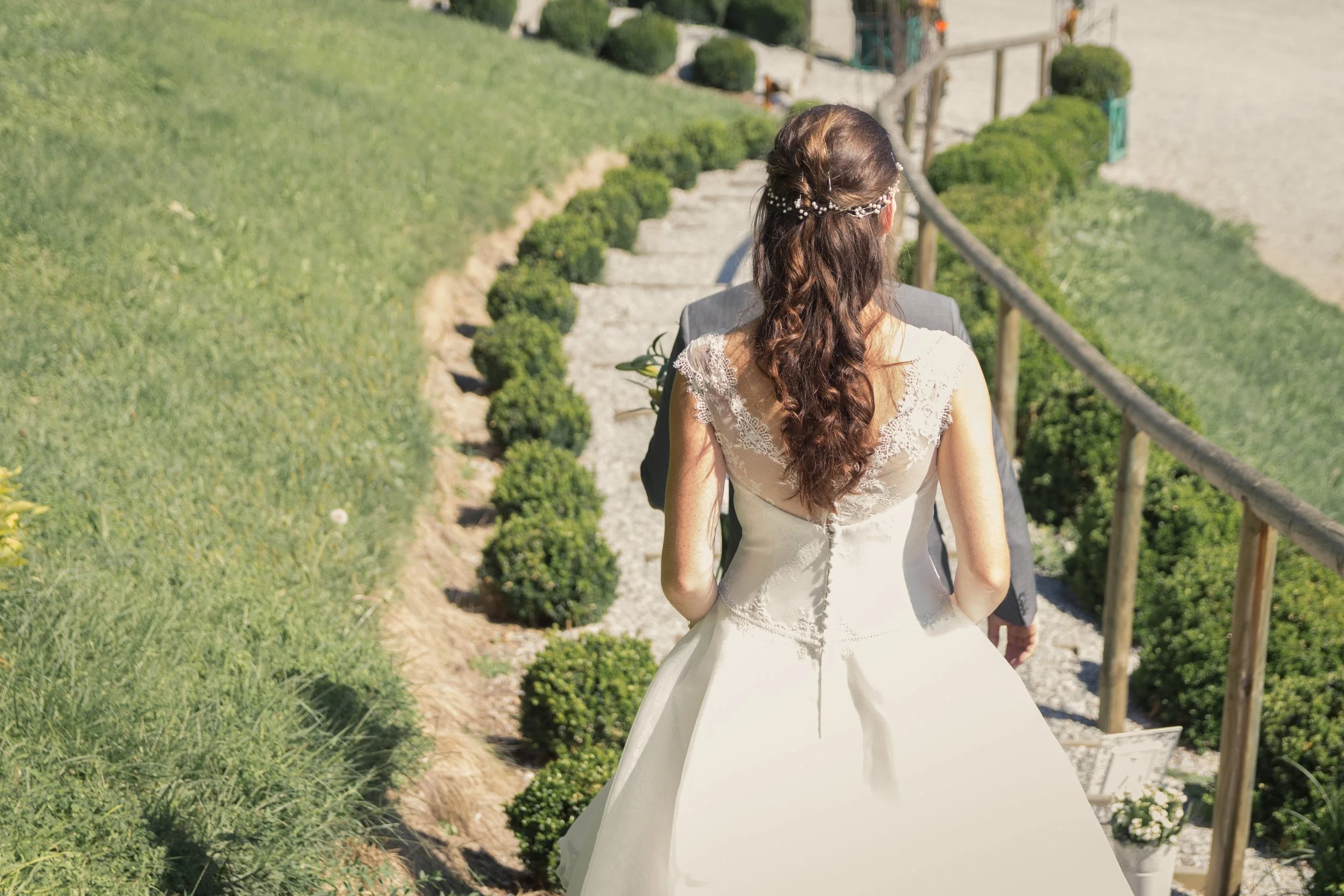 Braut in weißem Brautkleid mit Locken und Haarkränzchen, die einen Mann auf einer Gartenstiege begleitet.