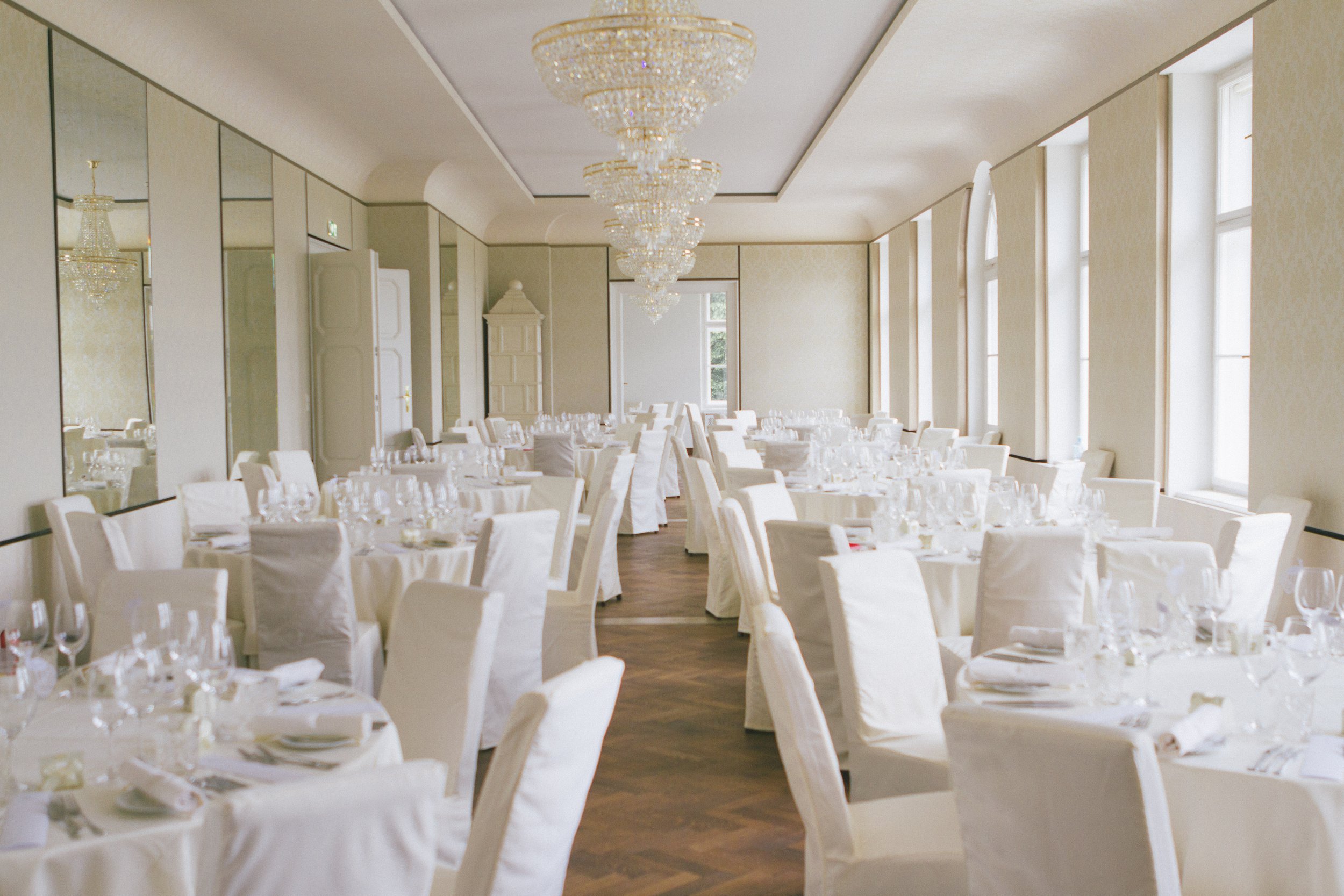 Ein festlich eingedeckter Saal mit weißen Tischen und Stühlen, mehreren Kristalllüstern an der Decke und großen Fenstern, die viel Tageslicht hereinlassen.