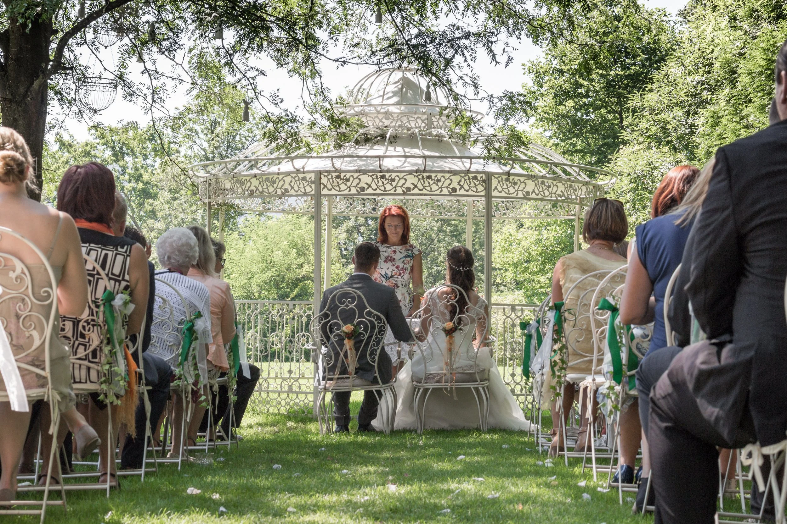 Eine Hochzeit im Freien mit Gästen, die auf Stühlen in einem Garten sitzen, während das Brautpaar vor einer weißen Gartenlaube sitzt und die Zeremonie stattfindet.