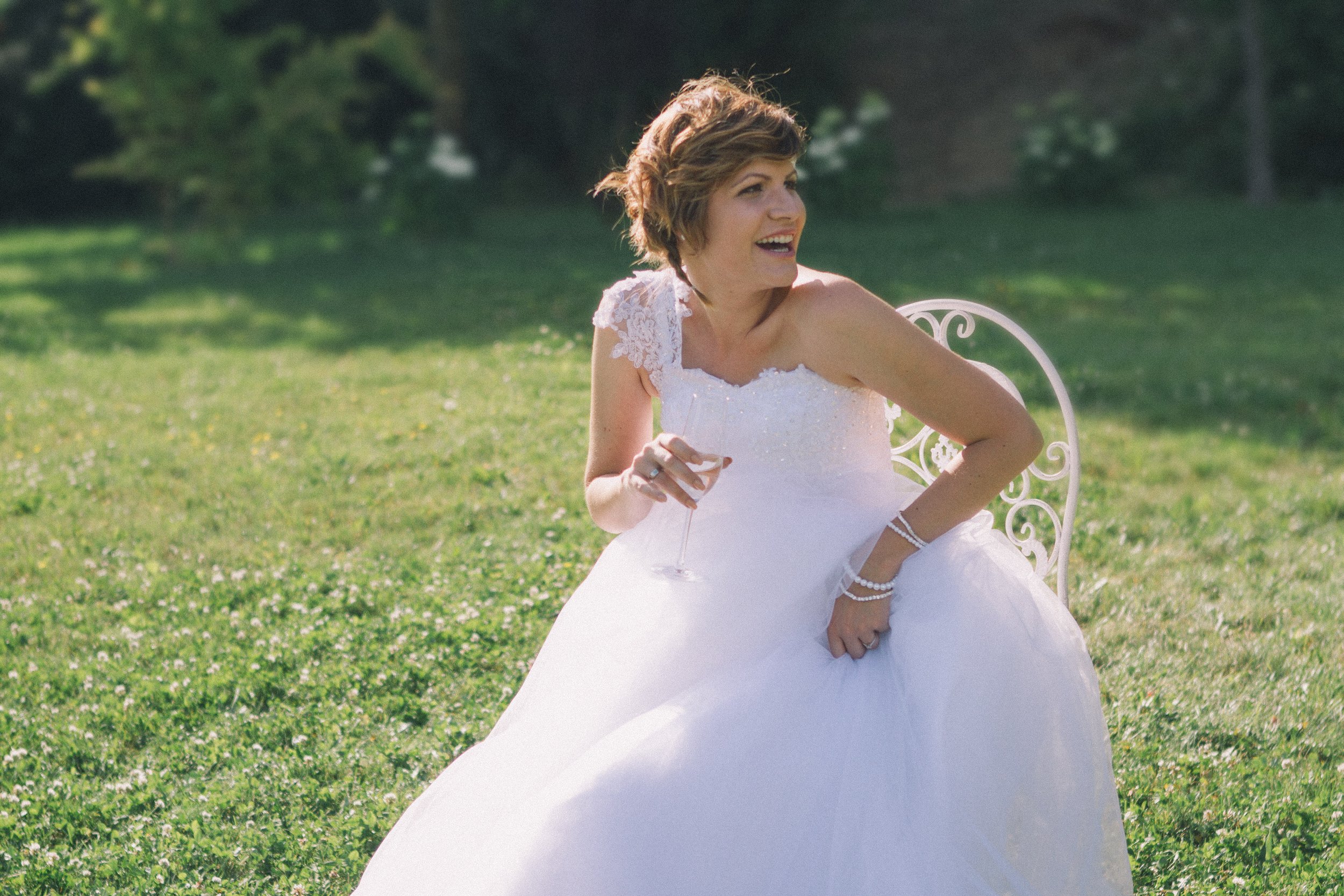 Eine lachende Frau in einem Hochzeitskleid sitzt im Garten auf einem weißen Stuhl, hält ein Sektglas in der Hand und ist umgeben von grünem Gras und Bäumen.