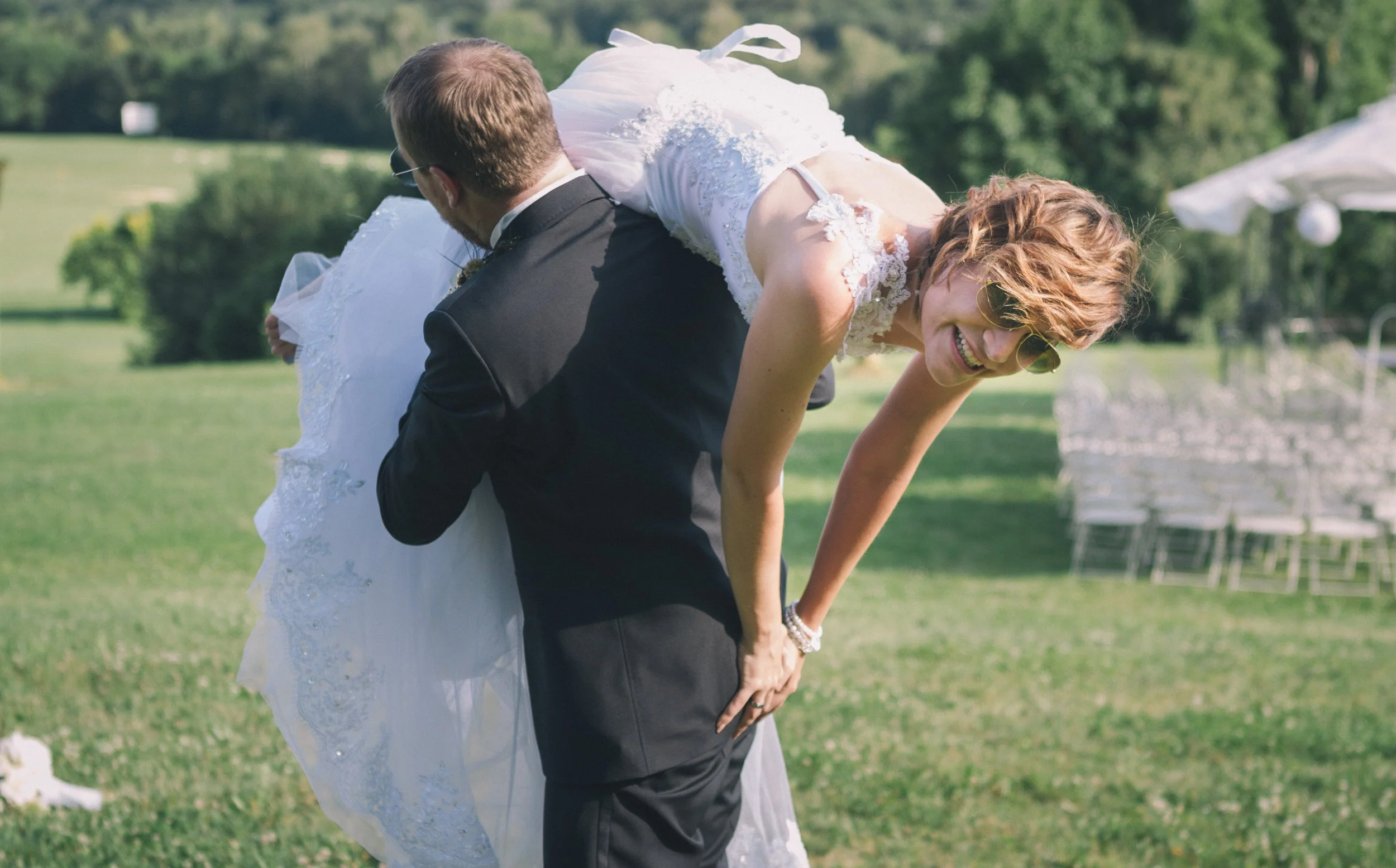 Ein Mann hebt eine lachende Frau bei einer Hochzeit im Freien hoch, beide tragen formelle Kleidung. Im Hintergrund sind Stühle und grüne Bäume zu sehen.