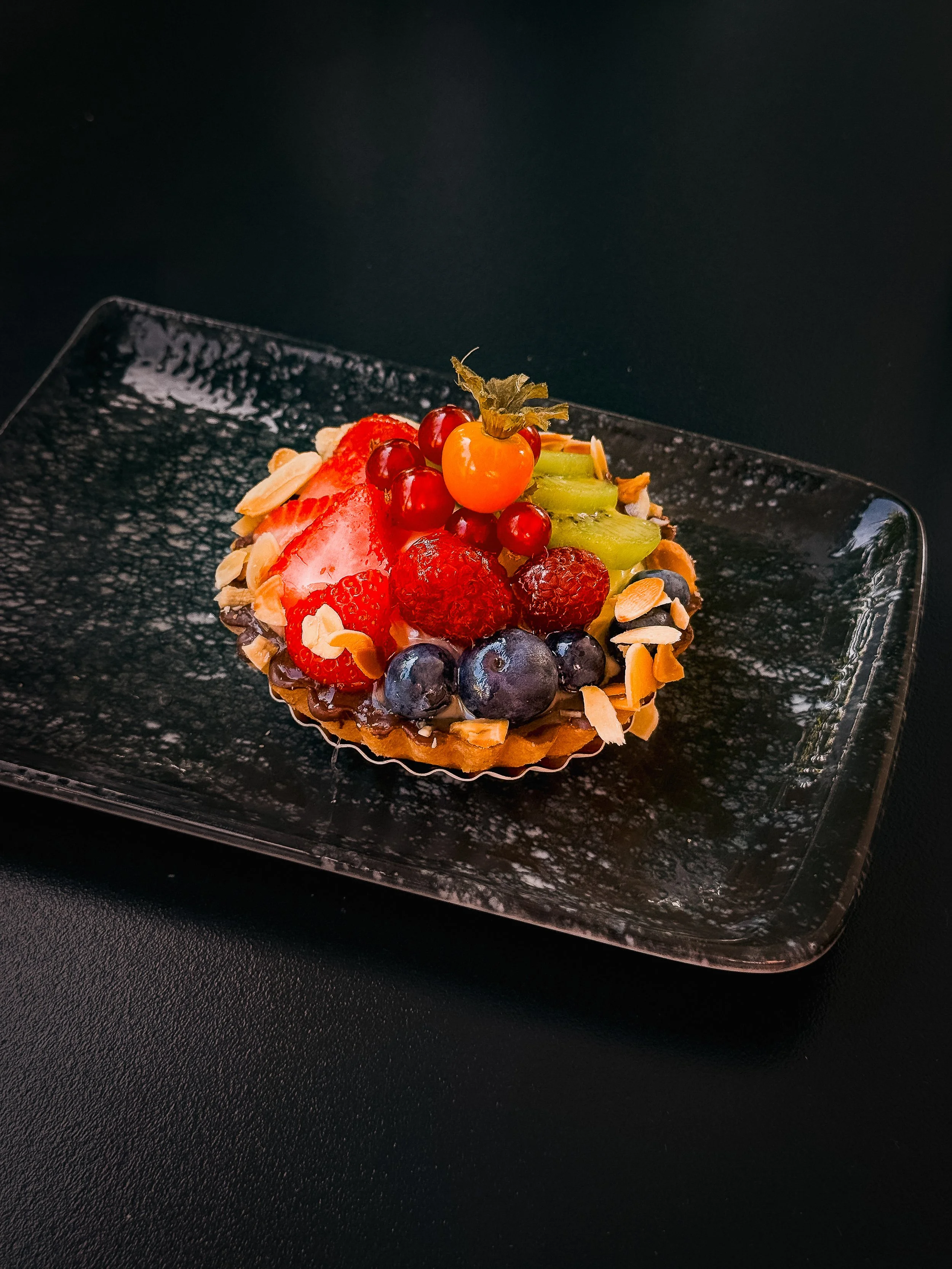 Obsttorte mit frischen Früchten wie Erdbeeren, Heidelbeeren, Grapefruit, Weintrauben, und einer kandierten Physalis, dekoriert mit Mandelblättchen, auf einem schwarzen Teller.