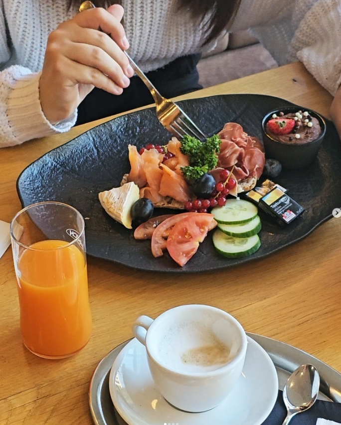 Frühstückstisch mit einem schwarzen Teller mit Antipasti, Gurkenscheiben, Tomatenscheiben, Käse, aufgeschnittene Zitrone, Weintrauben, Kirschen, Wurst und Schokolade. Ein Glas Orangensaft, eine Tasse Cappuccino und ein Löffel auf dem Tablett.