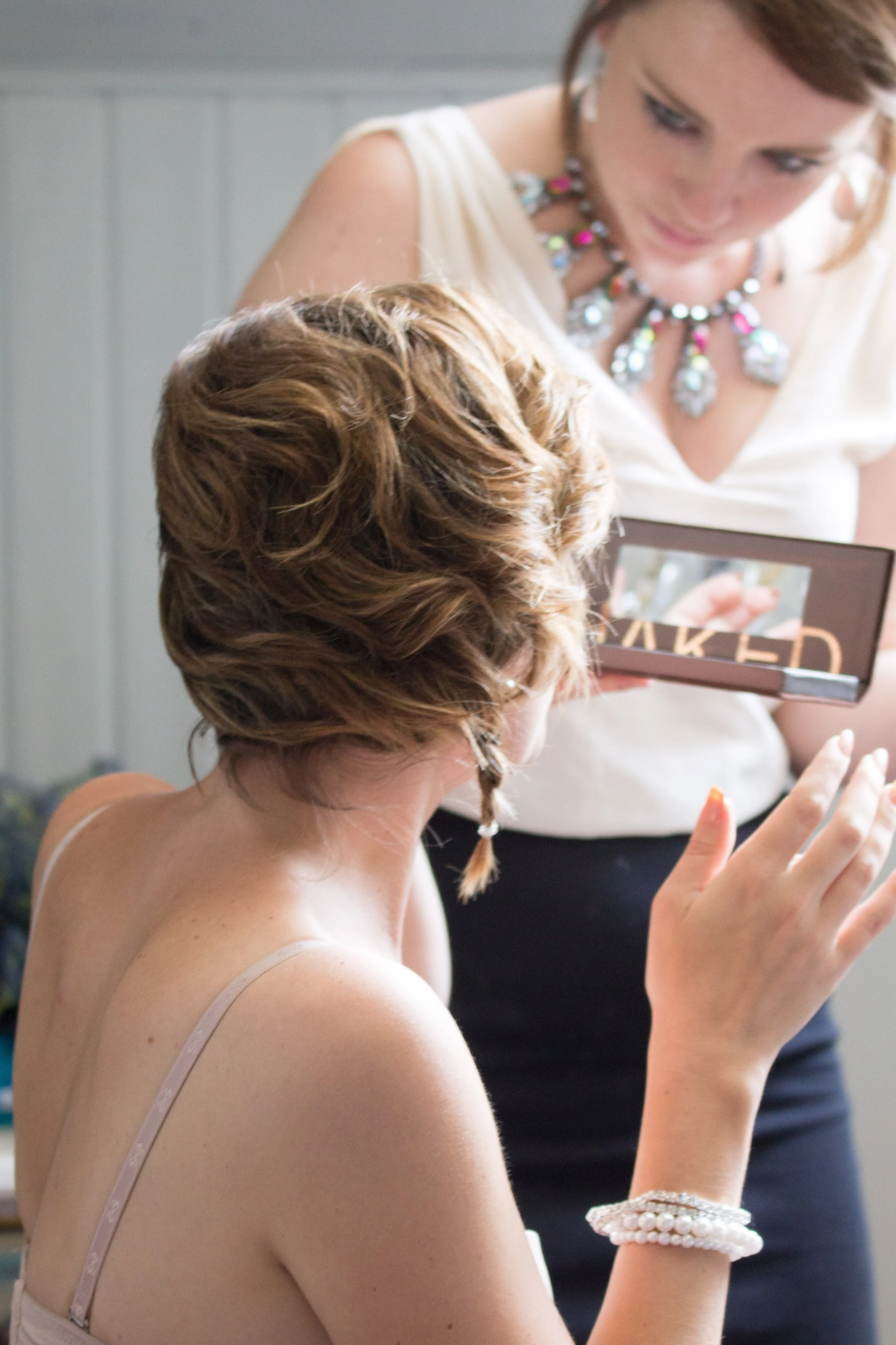 Eine junge Frau mit braunen Locken trägt ein weißes Oberteil, Schmuck und ein Armband aus Perlen, während sie mit einer anderen Person ein Makeup-Produkt betrachtet.