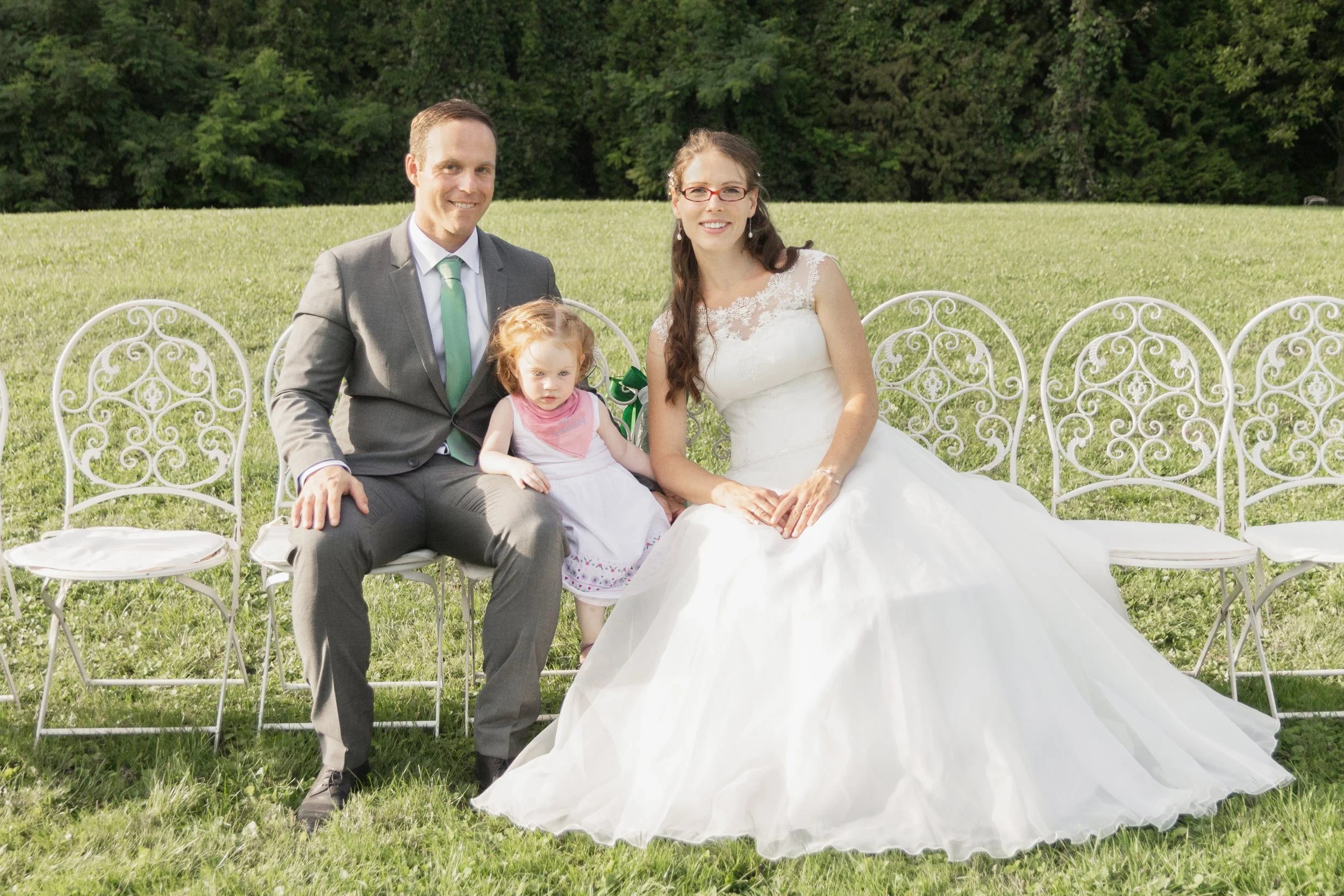 Ein Hochzeitsfoto von einer Frau in weißem Kleid, einem Mann in Anzug und einem kleinen Mädchen, die auf weißen Gartenmöbeln im Freien sitzen, mit grüner Wiese und Bäumen im Hintergrund.