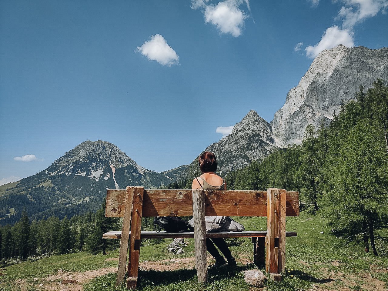 Eine Frau sitzt auf einer Holzbänke im Grünen, umgeben von Bergen und Bäumen, bei schönem Wetter mit blauen Himmel und Wolken.