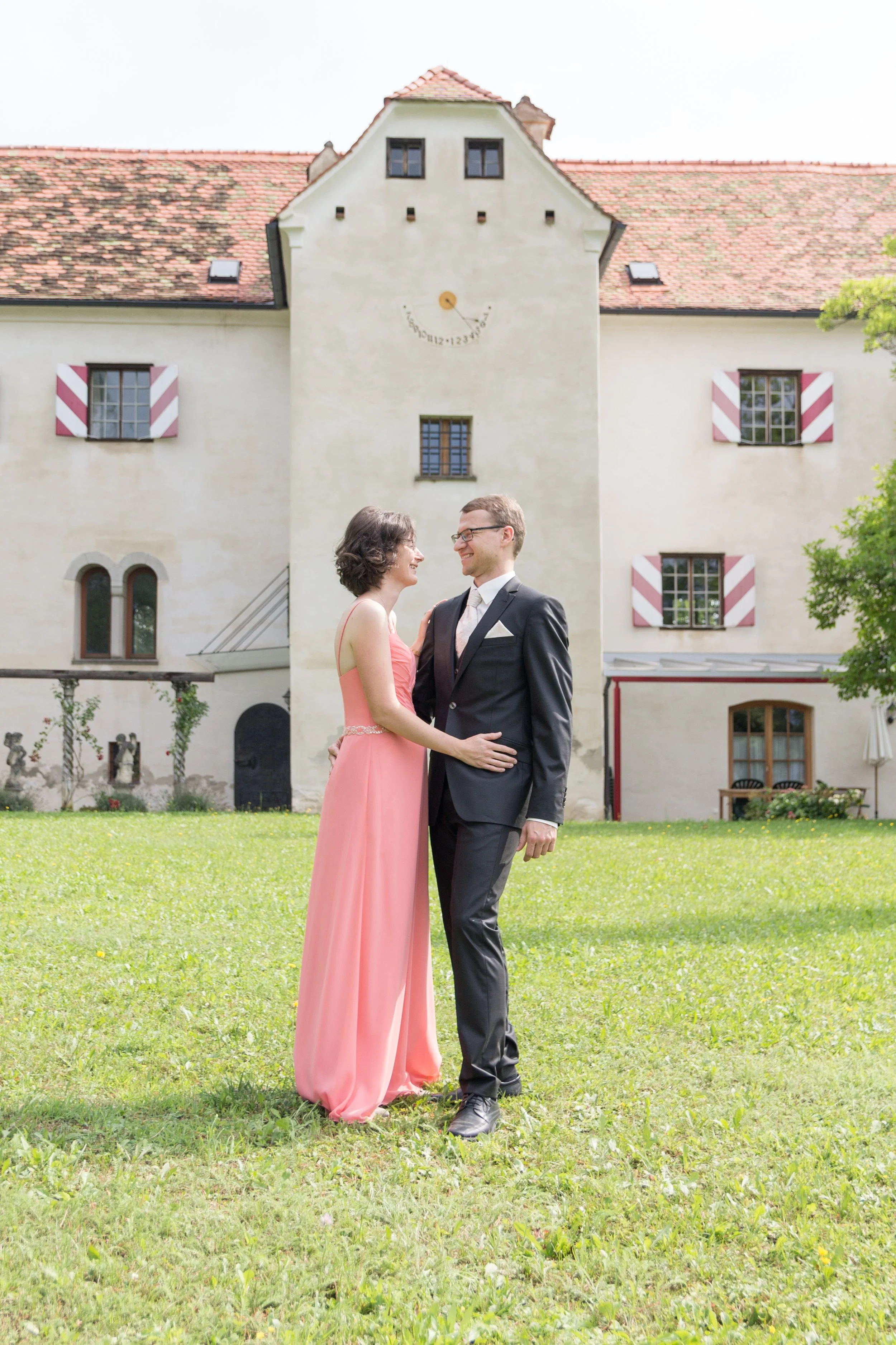 Ein Paar in formeller Kleidung, ein Mann in Anzug und eine Frau in einem langen pinken Kleid, stehen sich auf einer Wiese vor einem alten Gebäude mit roten Dachziegeln und kleinen schwarzen Fenstern gegenüber.