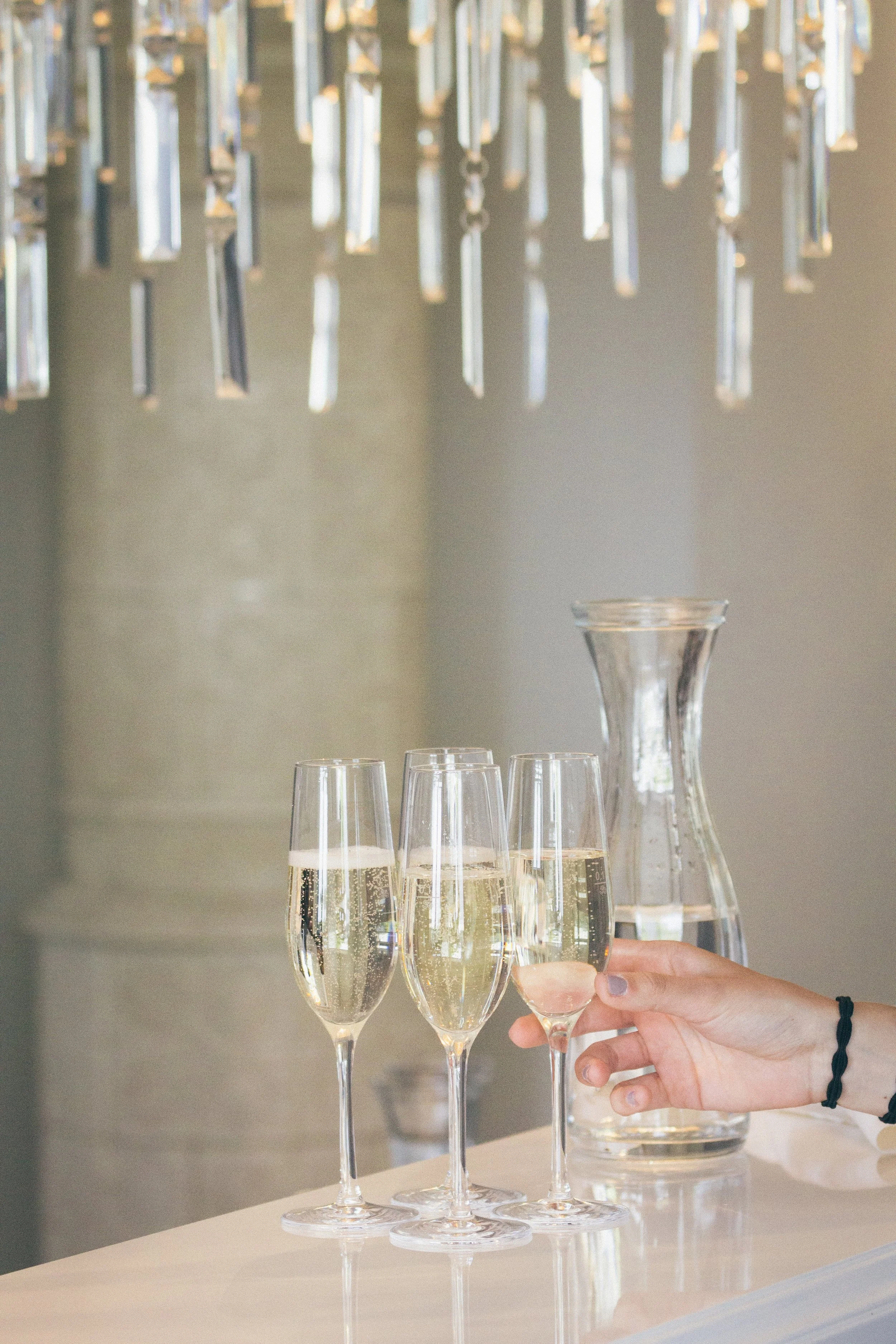 Person, die ein Glas Champagner hält, während mehrere andere Gläser und eine Wasser Karaffe auf einem Tisch stehen, mit einer dekorativen Hängelampe im Hintergrund.