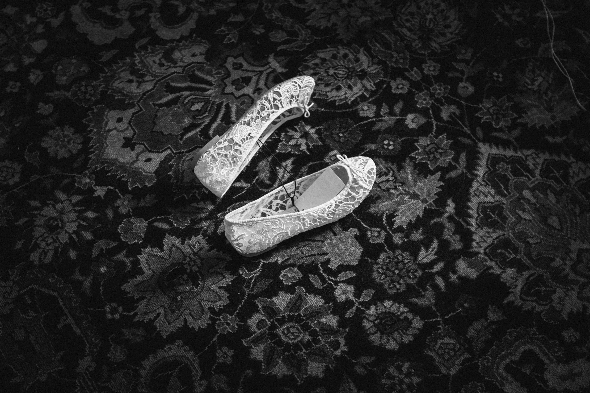 Zwei weiße, spitzenbesetzte Damenschuhe liegen auf einem schwarzen, floral gemusterten Teppich.