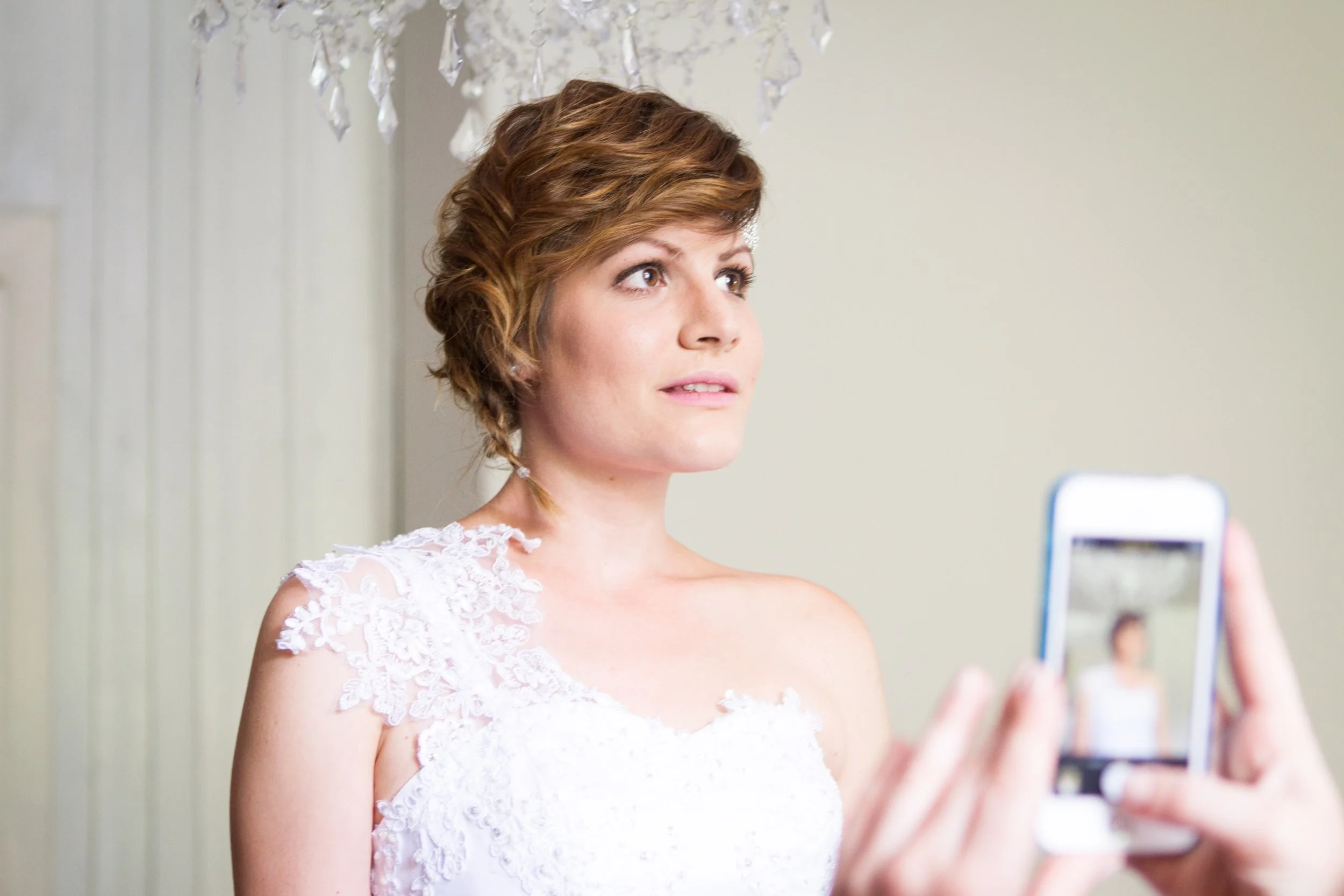 Braut macht ein Selfie mit einem Smartphone, während sie ein helles, lockiges Haarstyling trägt, in einem Raum mit neutralen Wänden und einer Kristalllampe.