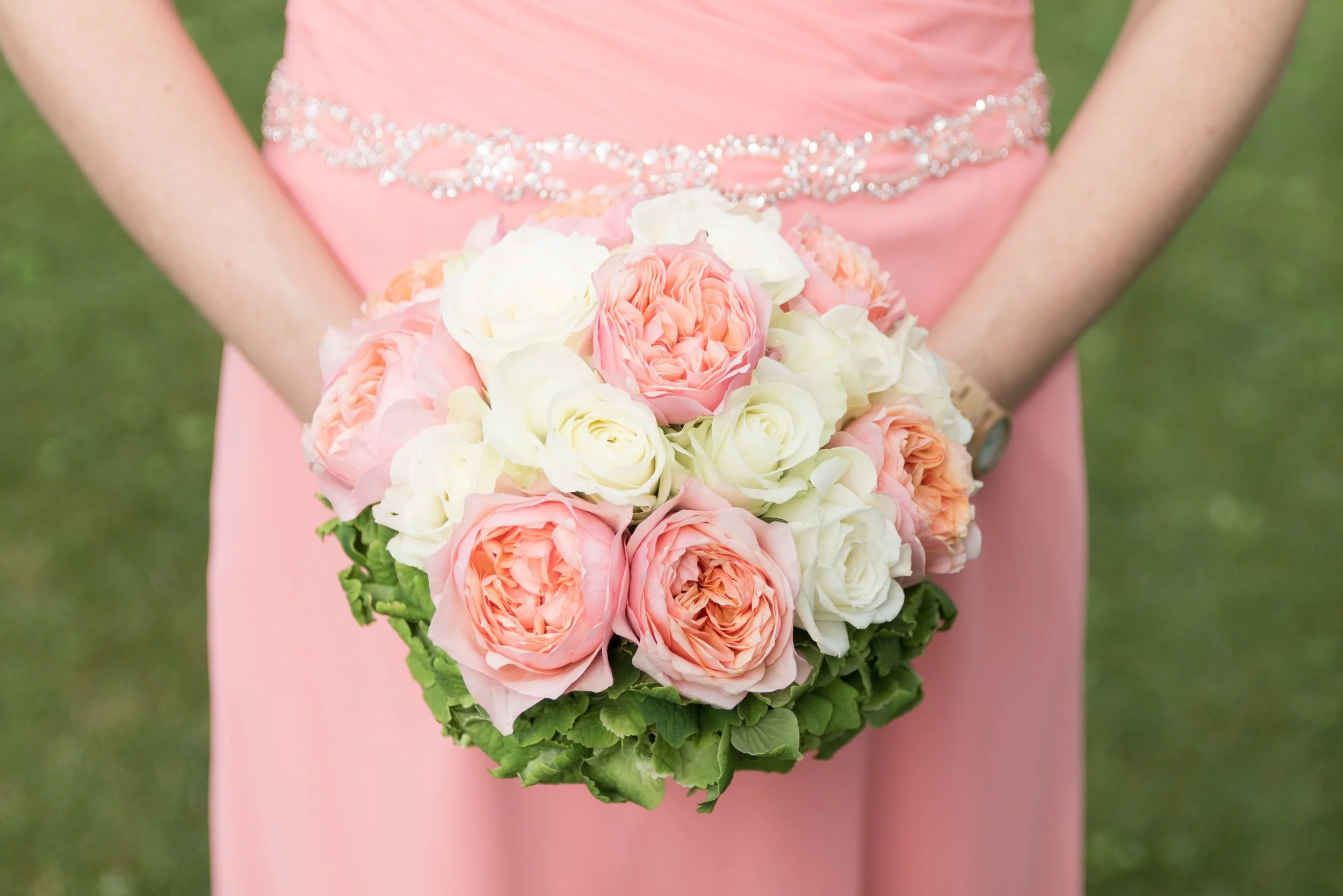 Person in einem pinken Kleid hält einen Blumenstrauß aus weißen, rosa und peachfarbenen Rosen vor sich, im Hintergrund ist grünes Gras sichtbar.