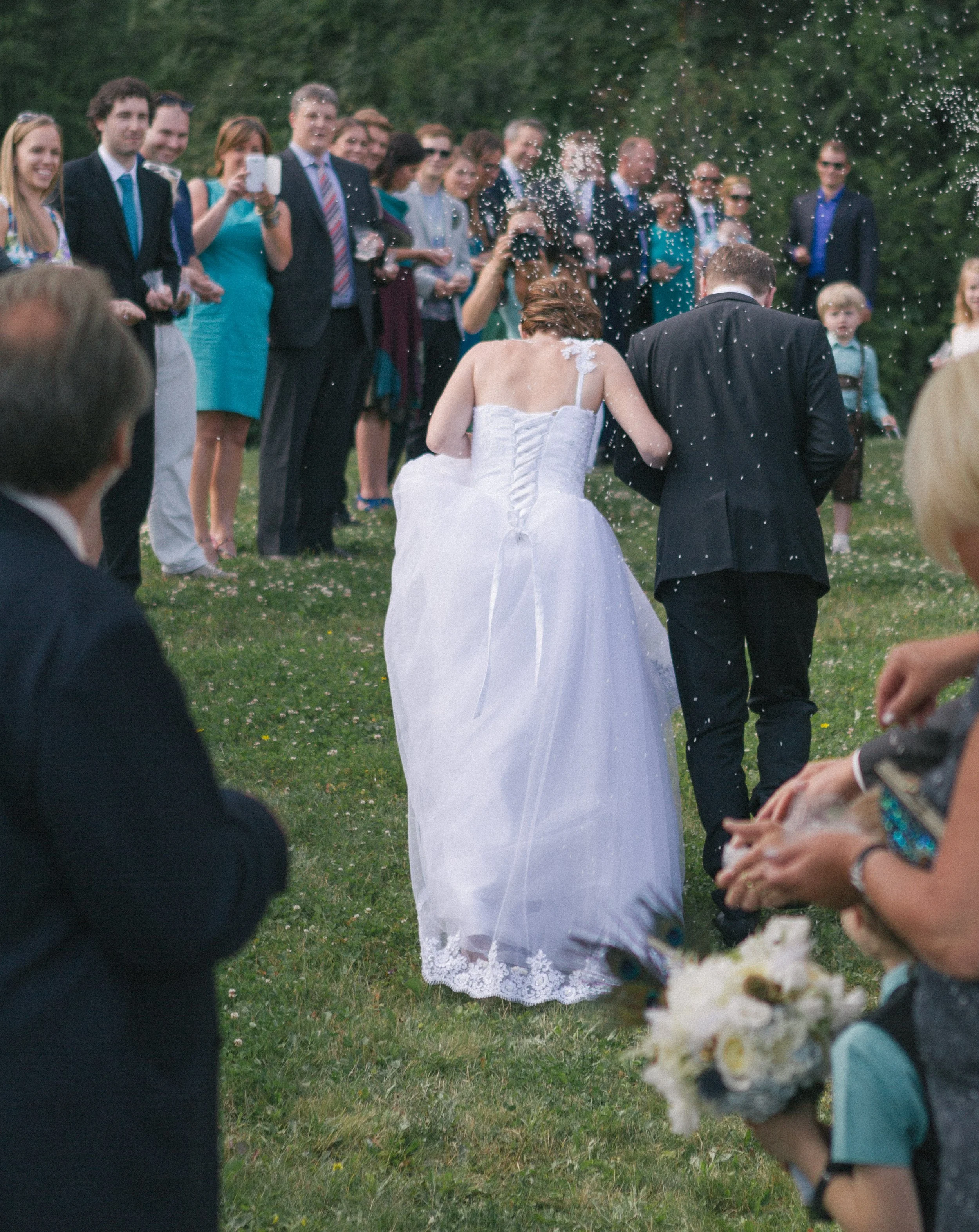 Hochzeitspaar mit Braut in weißem Kleid und Bräutigam in Anzug beten bei einer Zeremonie im Freien, während Gäste zuschauen und Wasser spritzt.