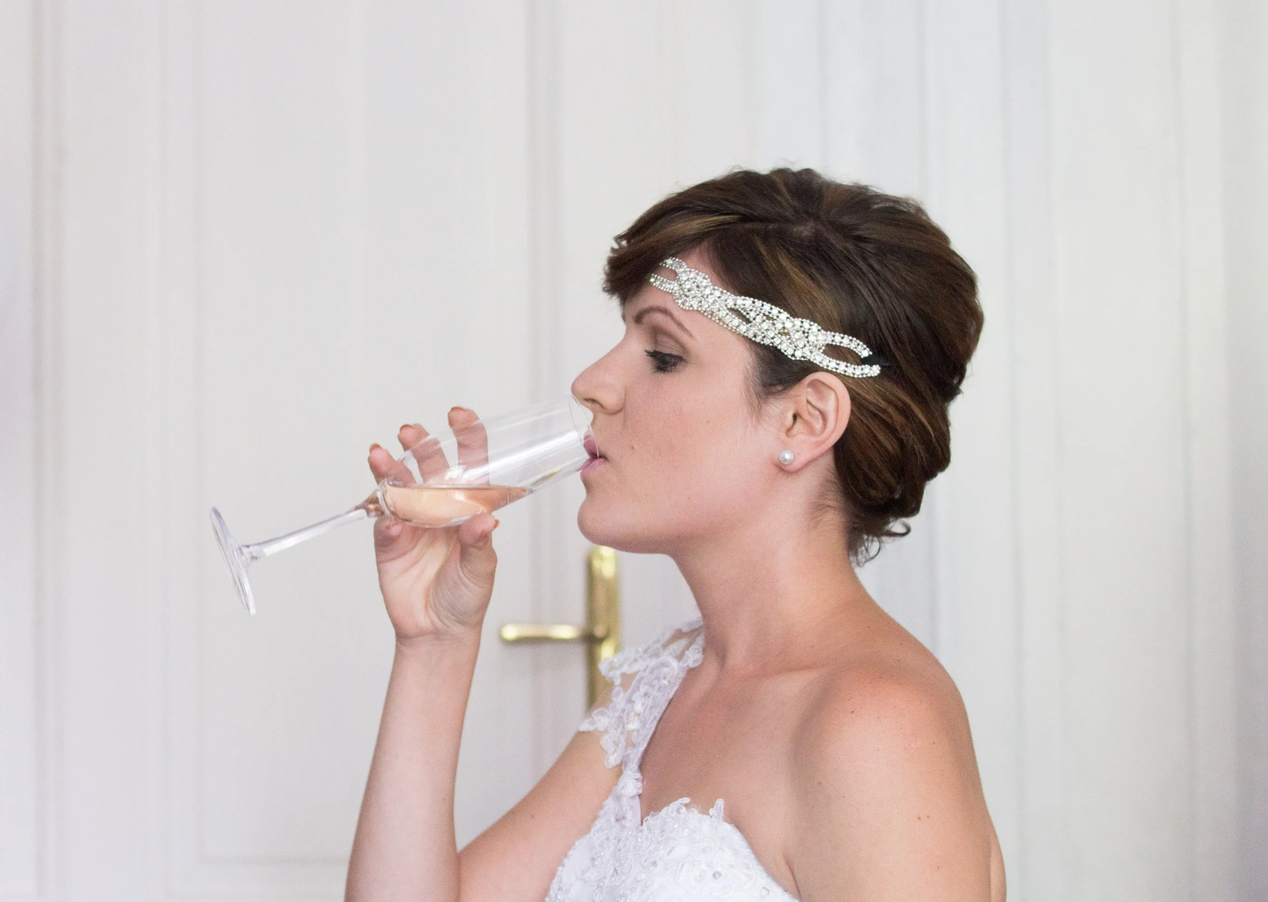 Eine Frau mit braunen kurzen Haaren, einer silbernen Stirnband, Perlenohrringen und einem weißen Spitzenkleid trinkt ein Glas Sekt.