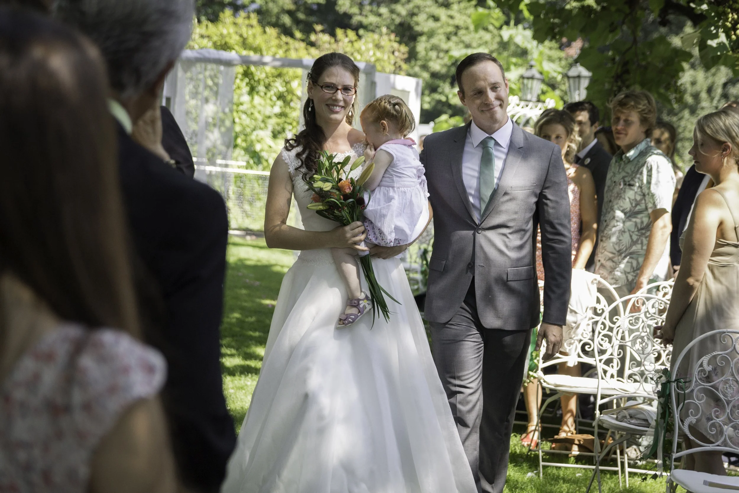 Braut hält ein Kind mit Blumenstrauß bei einer Hochzeit im Garten, umgeben von Gästen.