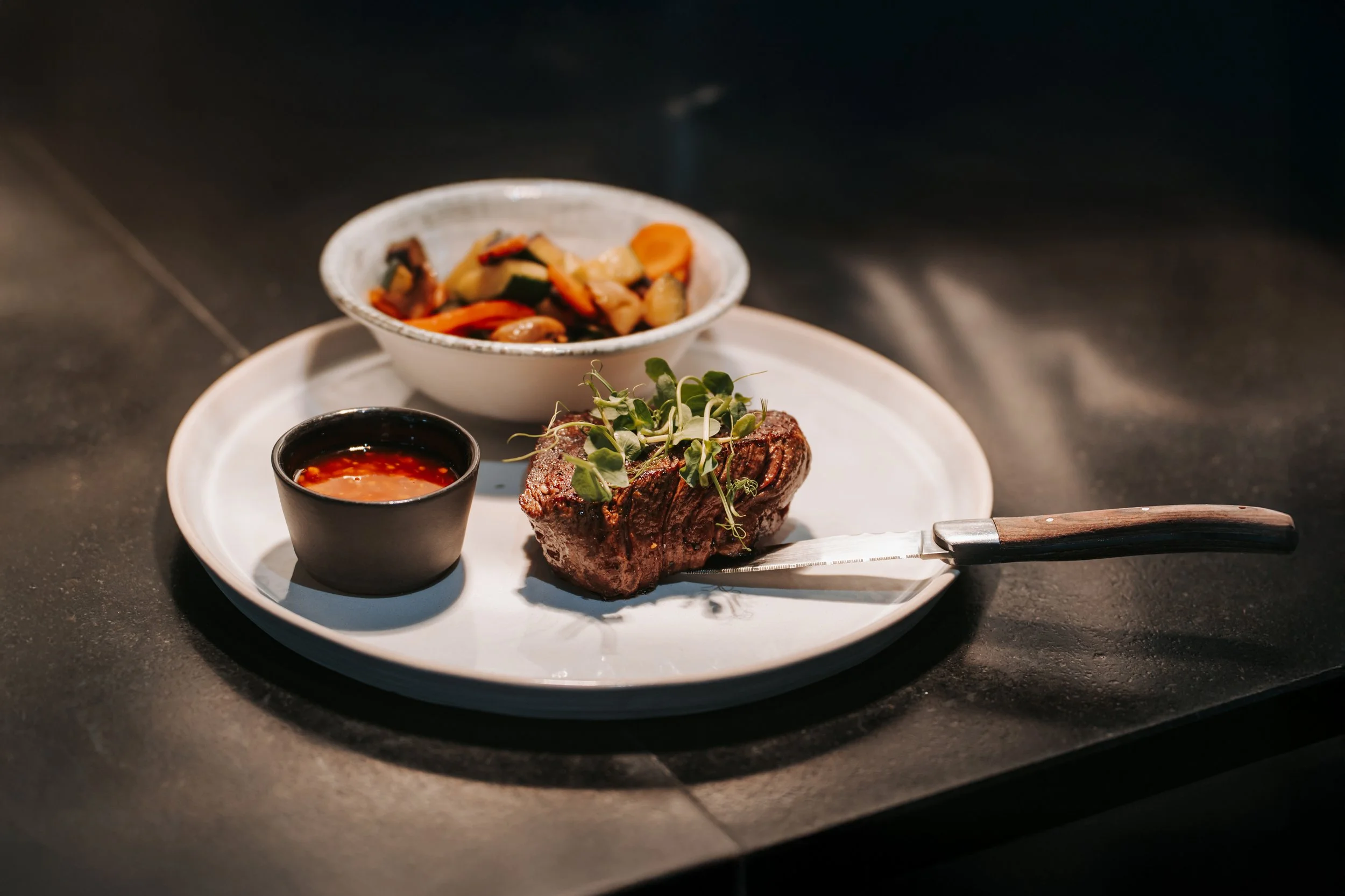 Ein Gericht mit einem Stück gebratenem Fleisch, garniert mit microgreens, dazu eine kleine Schüssel mit rotem Soßen und eine Schüssel mit gebratenen Gemüse im Hintergrund.