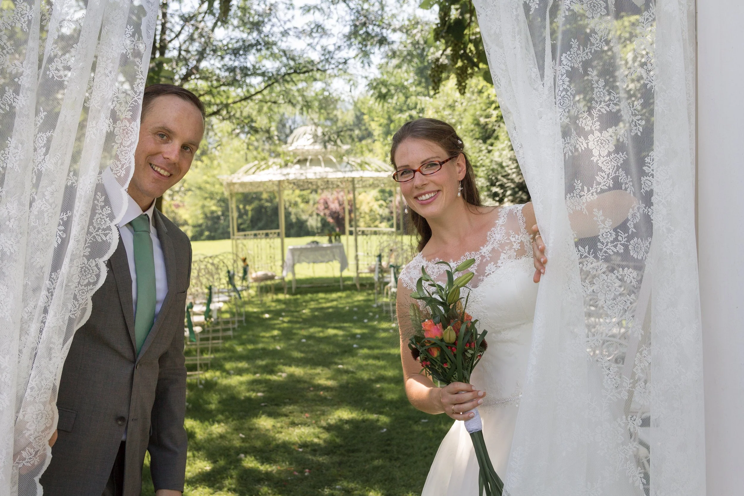 Ein lachendes Brautpaar steht im Garten, während sie durch Gardinen in einem weißen Pavillon schauen. Die Braut hält einen Blumenstrauß in der Hand.