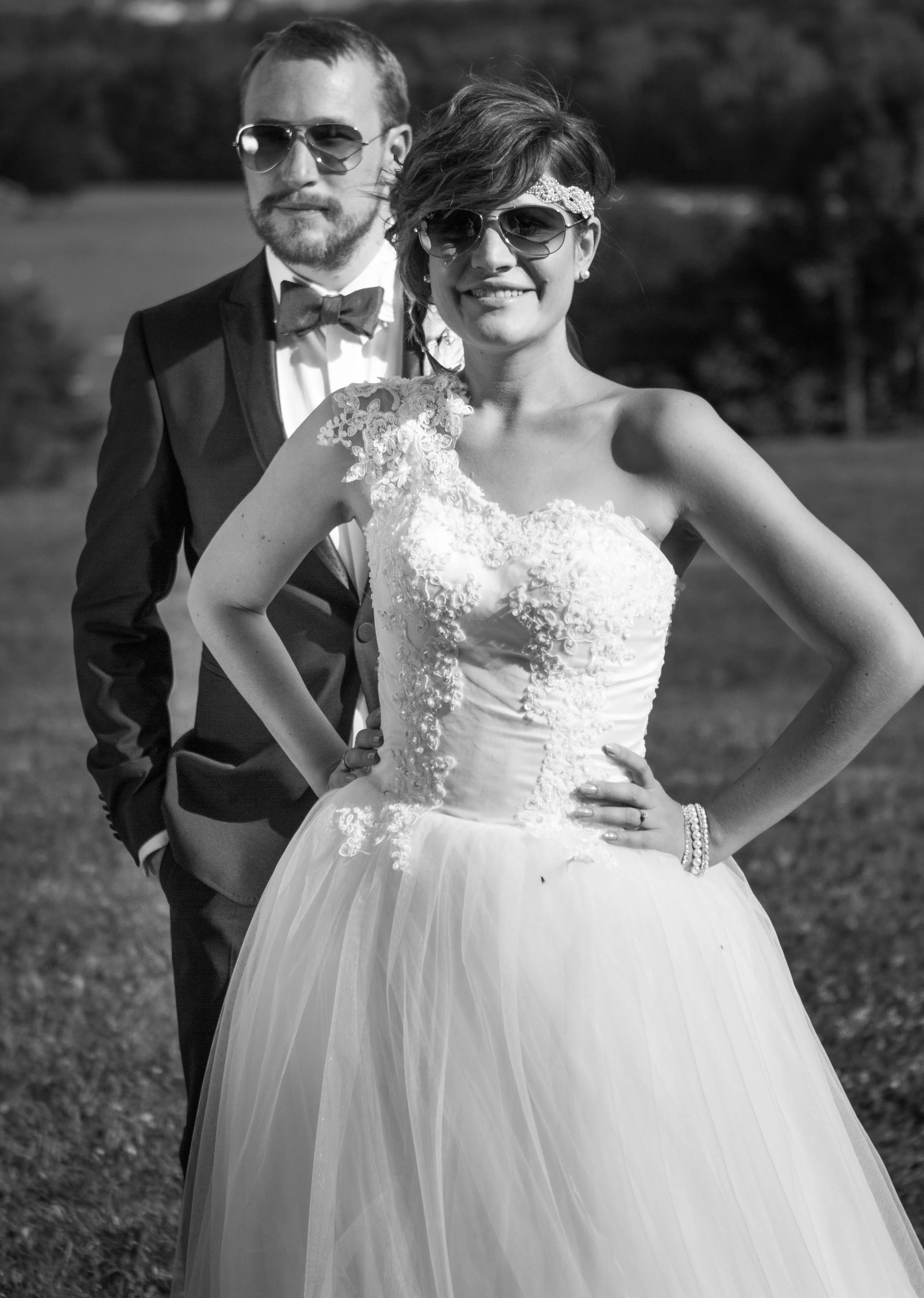 Ein schwarz-weißes Hochzeitspaar in formeller Kleidung, beide tragen Sonnenbrillen, draußen mit einem unscharfen Hintergrund von Bäumen und Natur.
