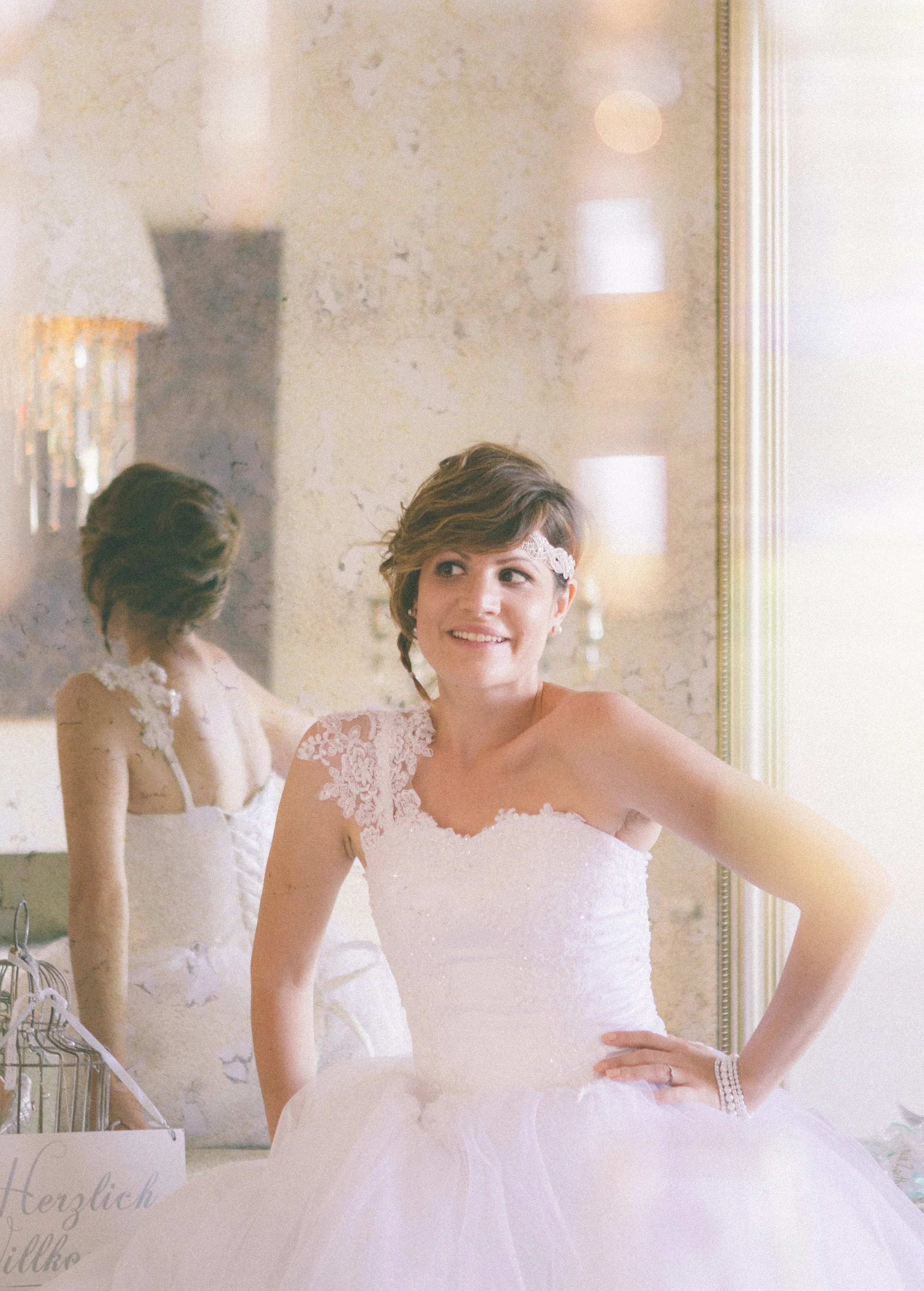 Junge Frau in Brautkleid schaut in einen Spiegel, trägt ein Haarband und hat einen glücklichen Ausdruck.