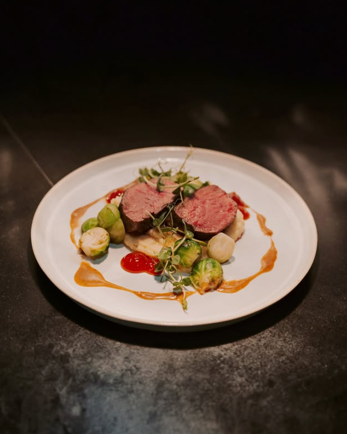 Auf einem weißen Teller liegt ein aufgeschnittenes medium-rare Fleischstück, garniert mit grünen und weißen kleinen Gemüse und einer Sauce
