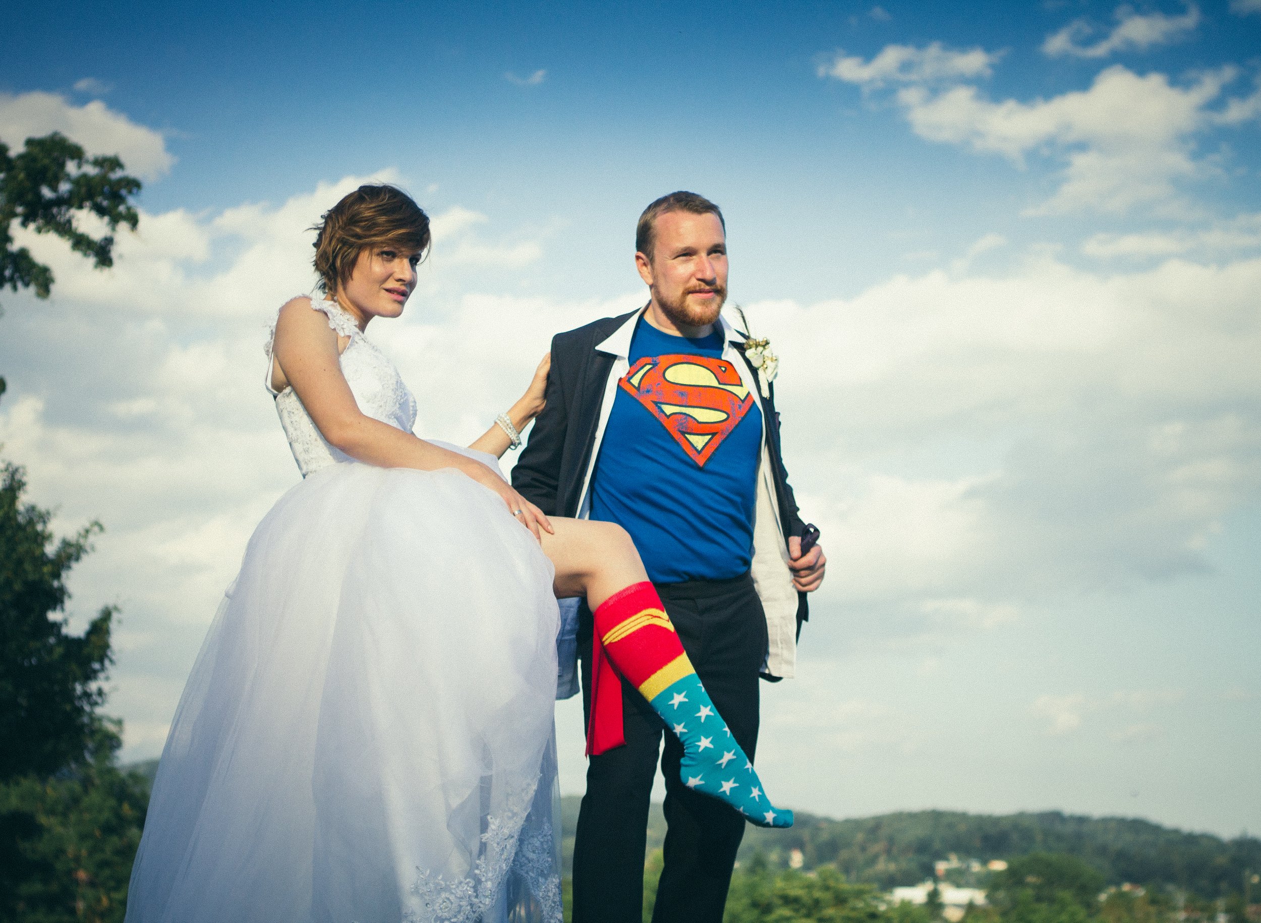 Ein Mann in einem Superman-T-Shirt trägt eine Frau in einem weißen Kleid mit Tutu, die scheinbar eine Braut ist, bei einem Fotoshooting im Freien unter einem blauen Himmel mit Wolken.