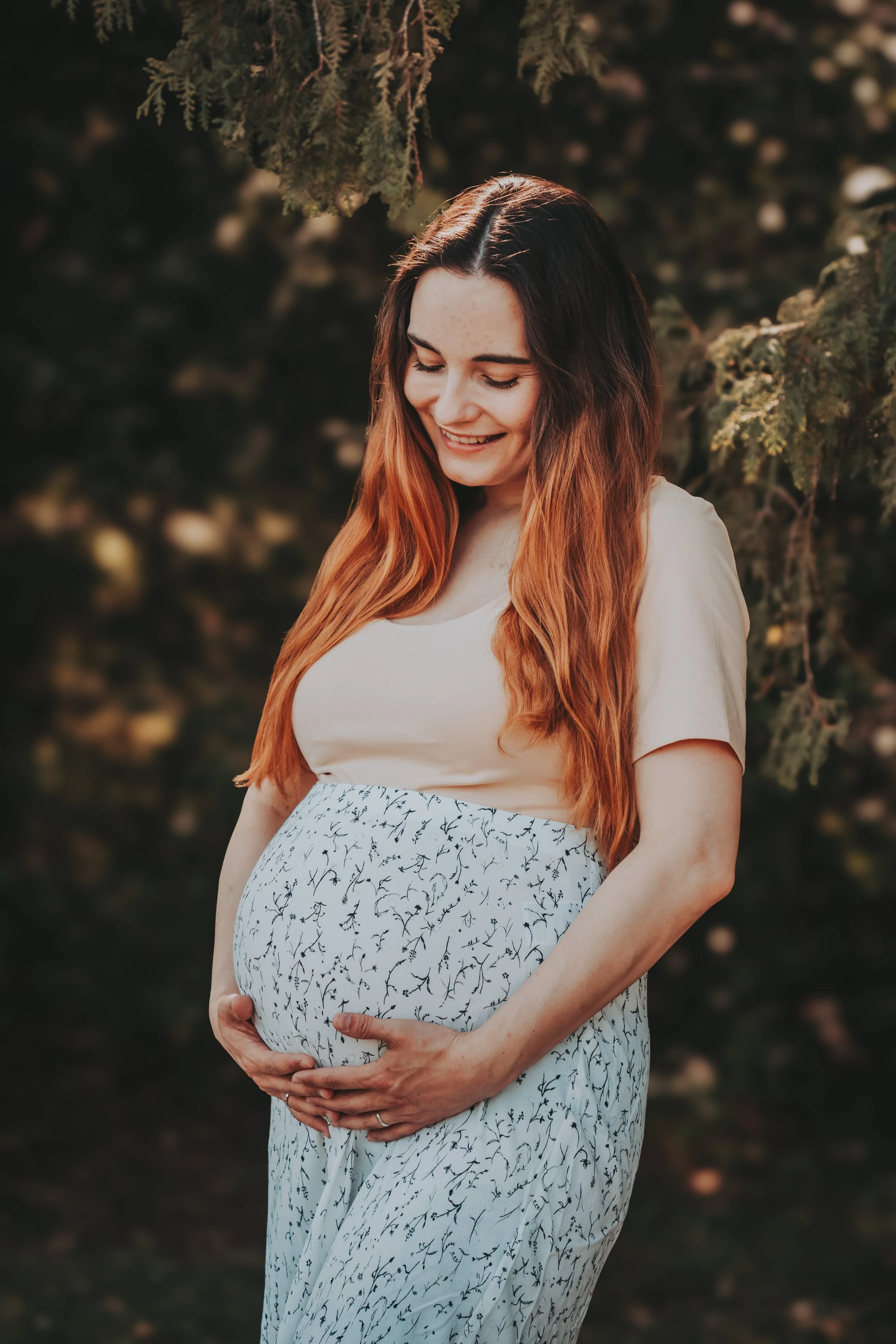 Eine schwangere Frau mit langen braunen Haaren, die vorsichtig ihren Babybauch hält, steht vor einem dunklen, grünen Hintergrund, lächelt und schaut schräg nach unten.