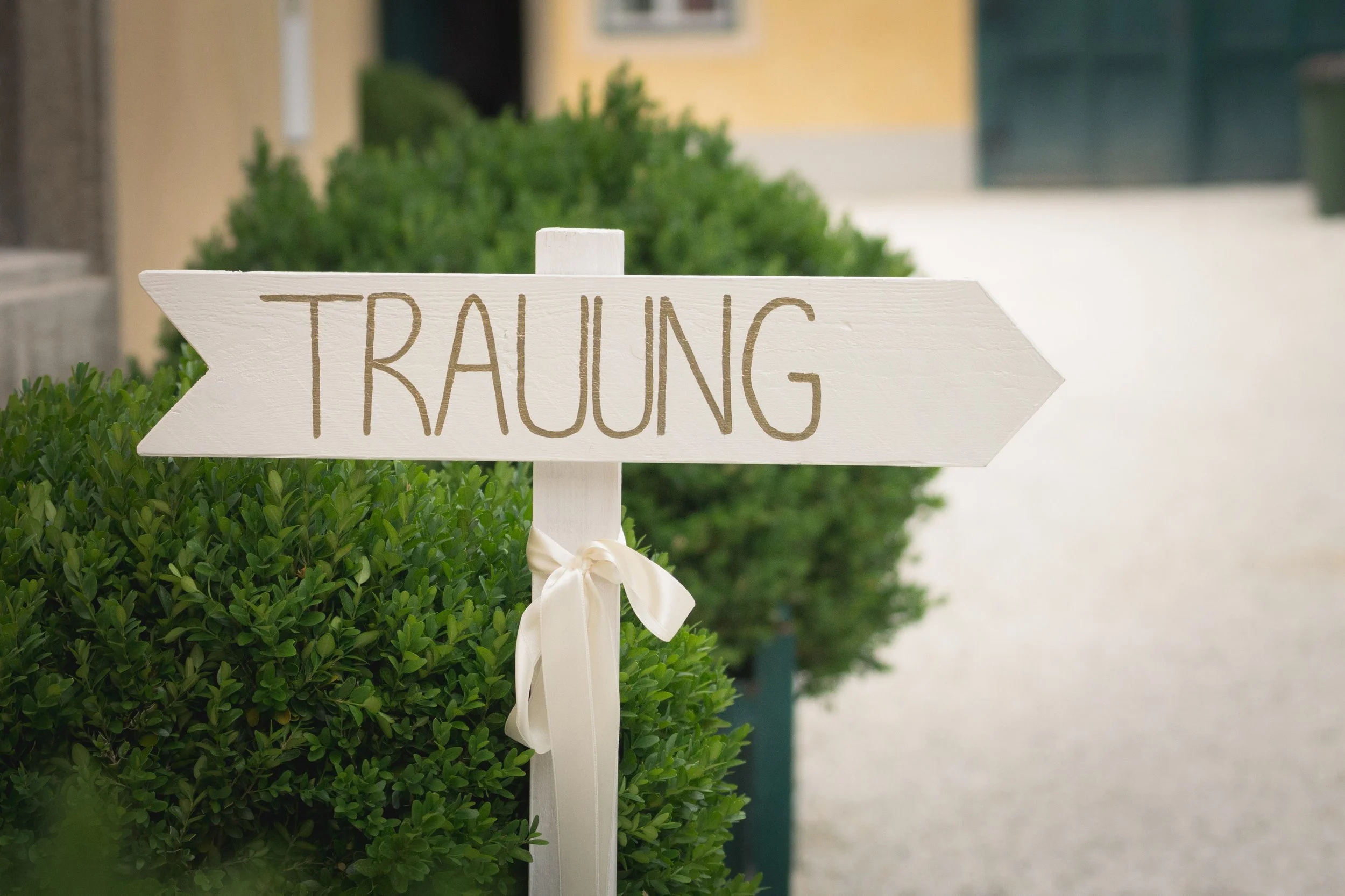Ein weißes Hinweisschild mit der Aufschrift 'TRaUNG' ist in einem Garten mit grünen Sträuchern aufgestellt.
