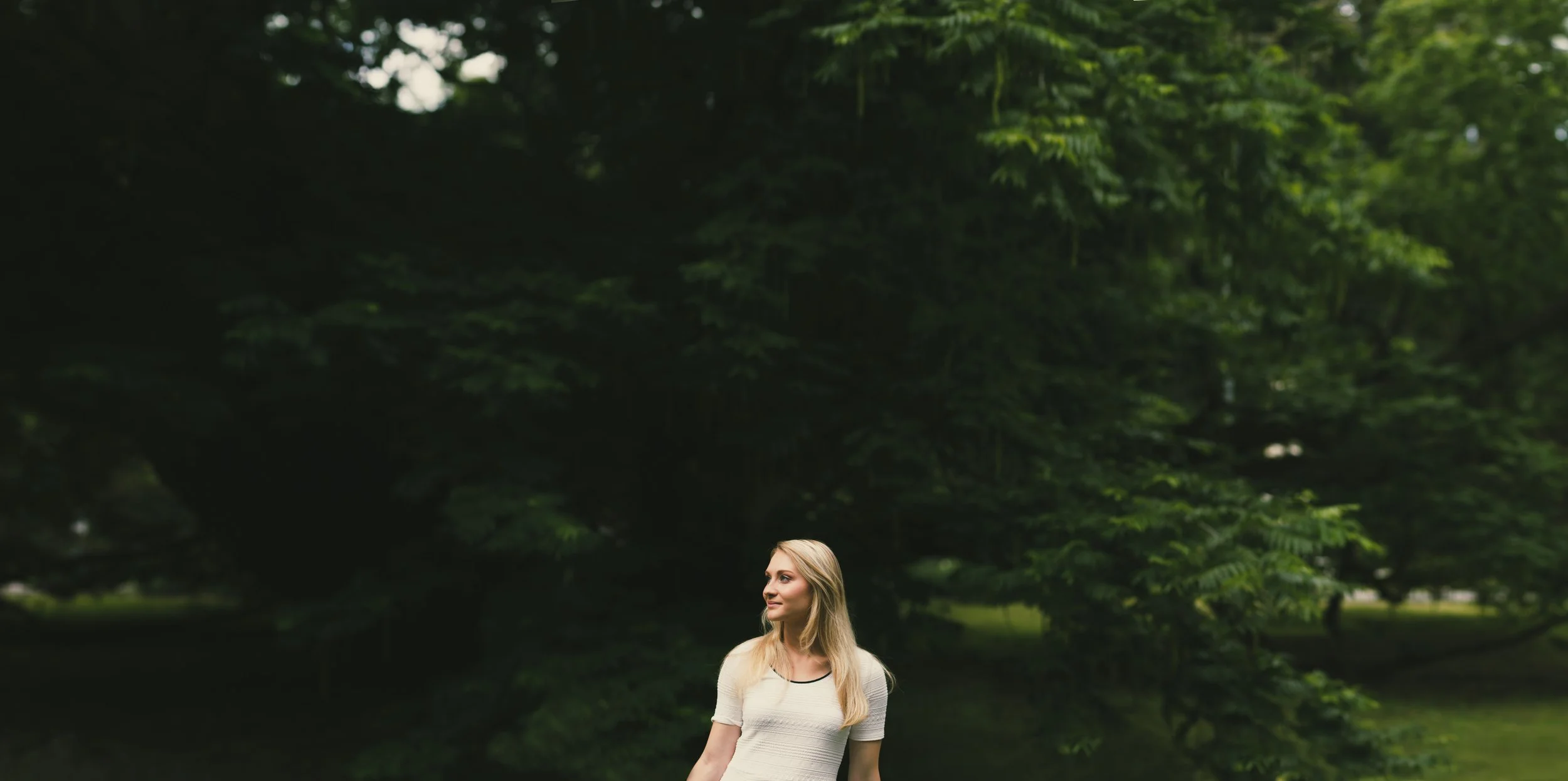 Eine blonde Frau steht in einem parkähnlichen Gebiet mit üppigem Grün und großen Bäumen im Hintergrund.