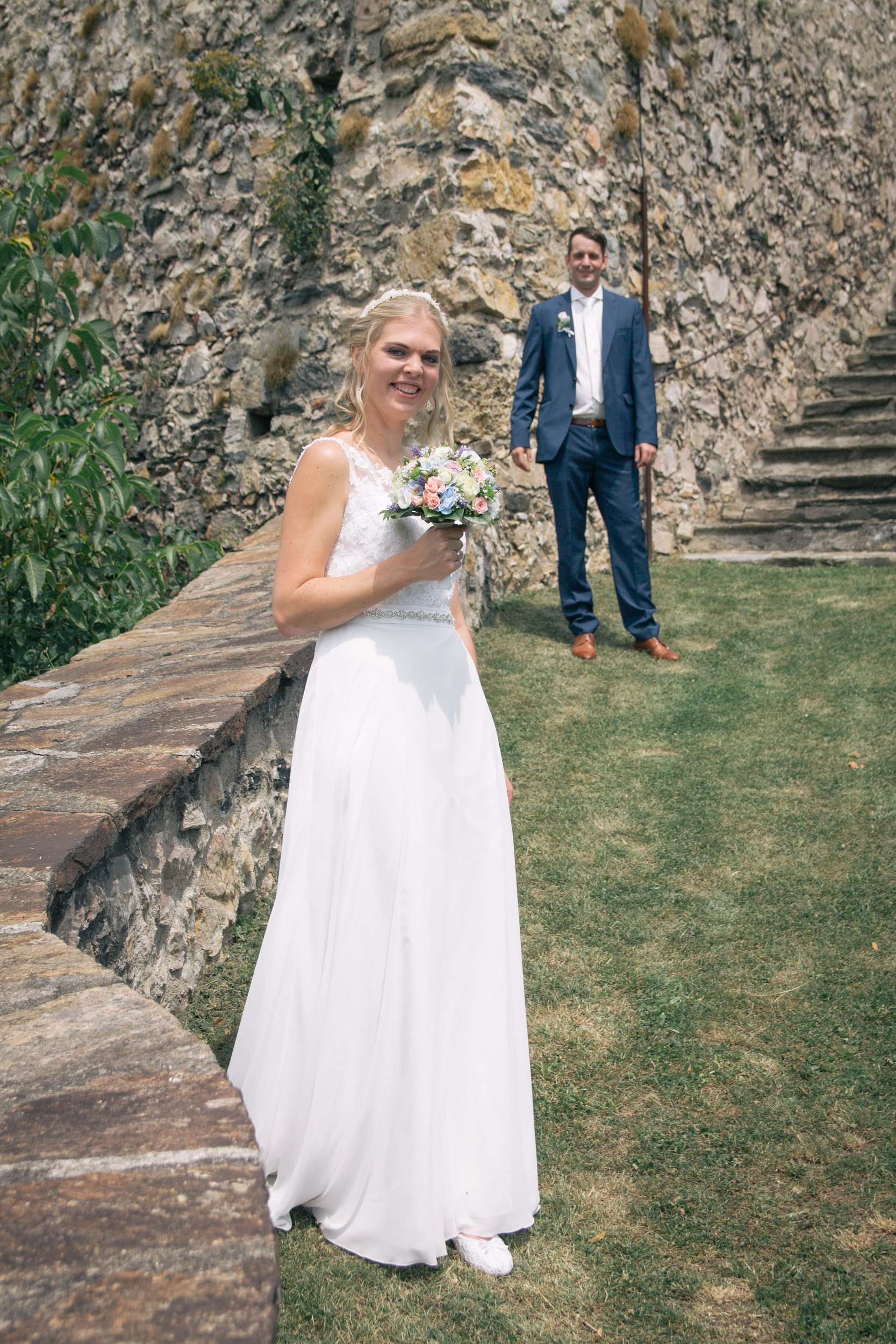 Braut im weißen Kleid mit Blumenstrauß lächelnd vor Steinmauer und grünem Garten, Bräutigam im blauen Anzug im Hintergrund
