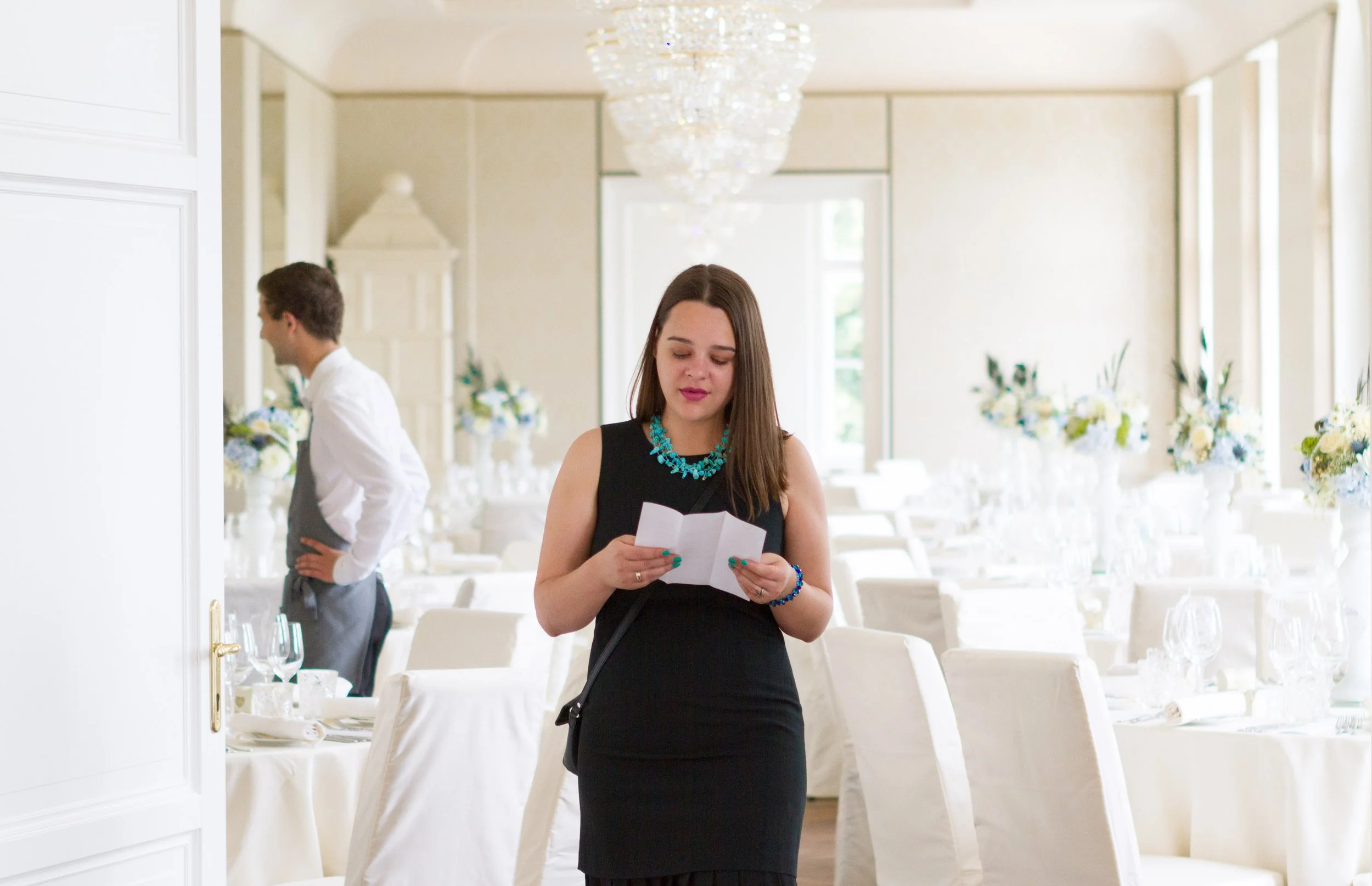 Eine junge Frau mit braunen Haaren, schwarzem Kleid und blauer Ketten liest in einem festlich dekorierten Raum eine Karte. Im Hintergrund sind weitere Menschen, Tische mit weißen Tischdecken und elegante Blumenarrangements zu sehen.