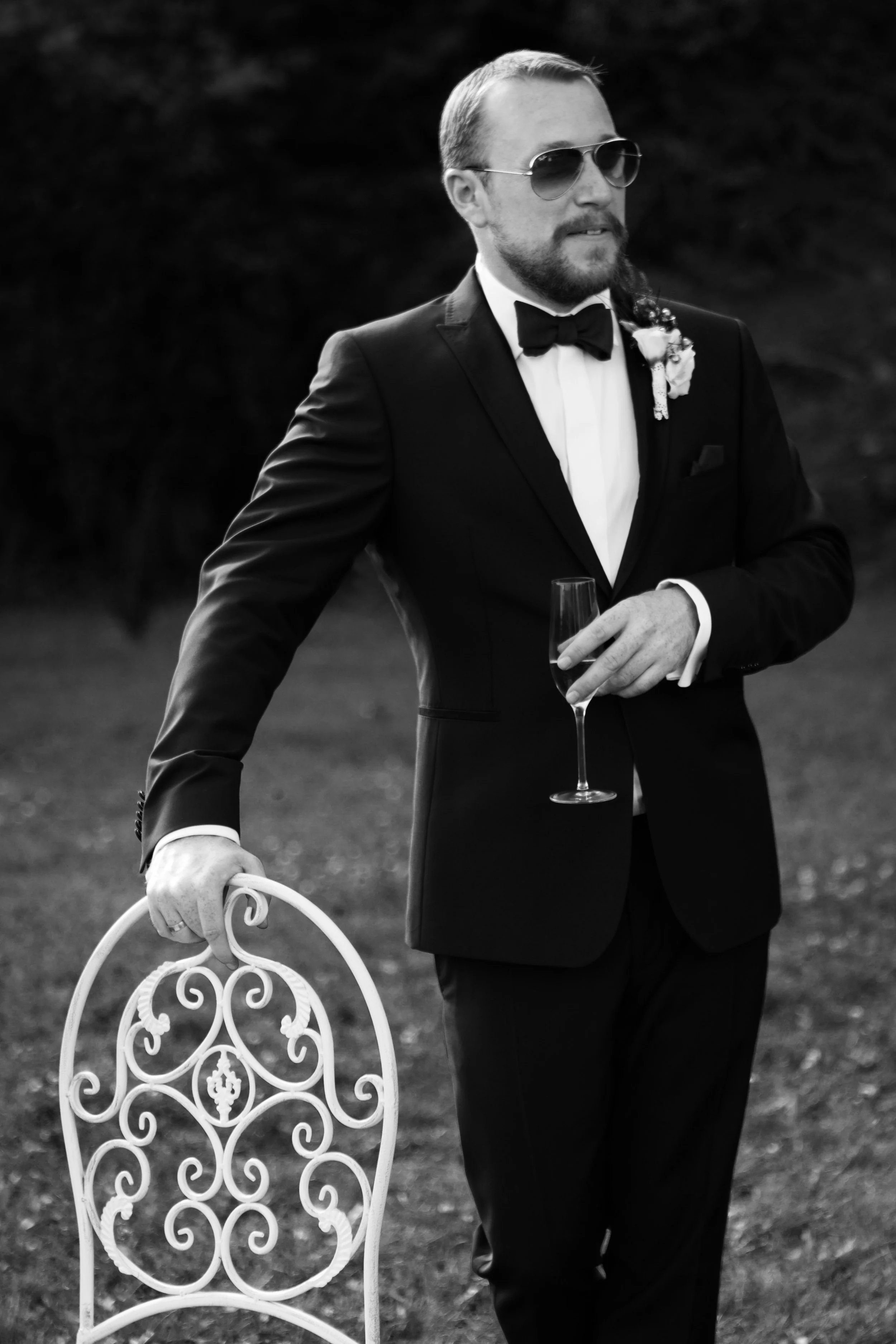 Schwarz-Weiß-Foto eines Bräutigams bei einer Hochzeit in formeller Kleidung mit Sonnenbrille, Fliege und Blumenanstecker, der eine Champagnerflöte hält, auf einem Gartenstuhl sitzend, im Hintergrund unscharfer Garten.