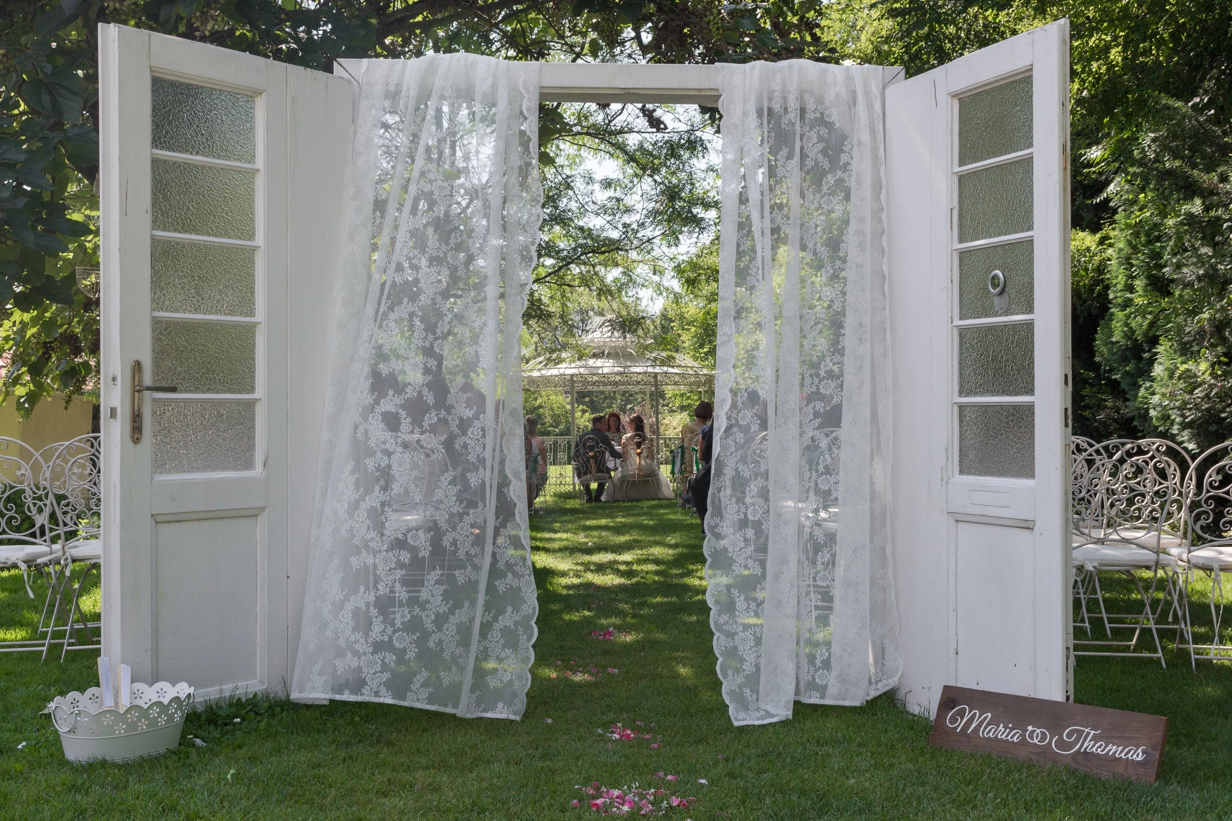 Ein Tor aus weißen Türen mit Glasfenstern, umgeben von weißen Gartenstühlen und Blumen, führt zu einem Garten mit einem Pavillon und einer Gruppe von Menschen bei einer Feier, vermutlich Hochzeit.