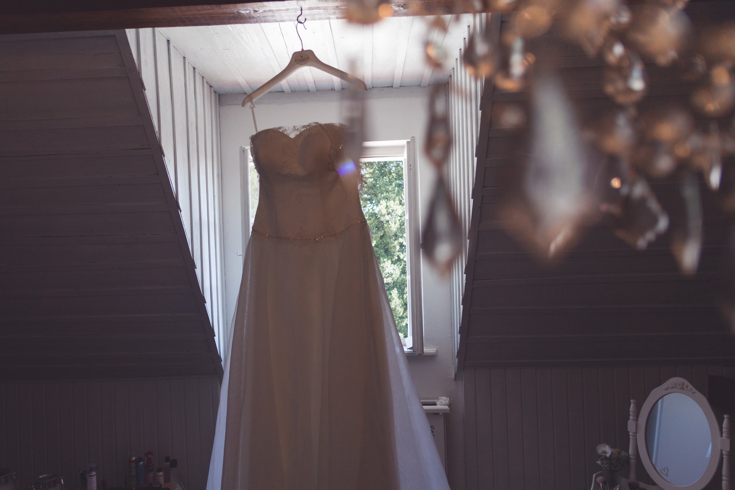 Ein Brautkleid hängt an einer weißen Kleiderstange vor einem Fenster, durch das Tageslicht scheint, während im Vordergrund eine warme herbstliche Dekoration zu sehen ist.