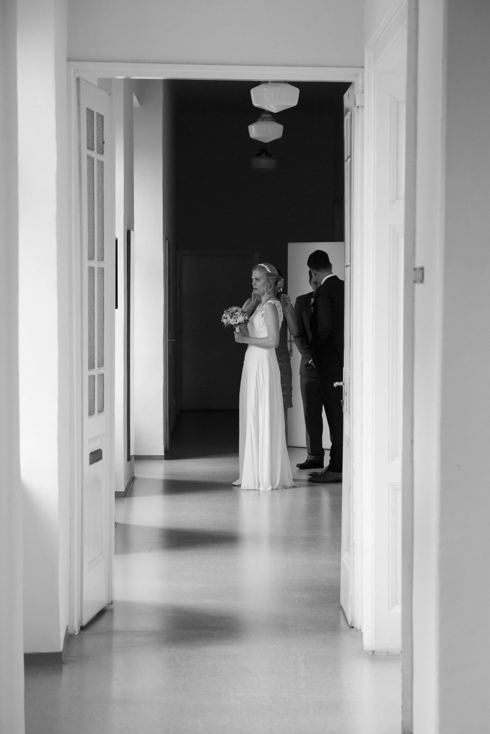 Eine Hochzeitsszene mit Braut in weißem Kleid und Veil, die in einem Flur steht, während der Bräutigam im Anzug mit dunkler Krawatte hinter ihr steht. Zwei weitere Personen sind im Hintergrund, alle im formellen Outfit. Die Szene ist schwarz-weiß.