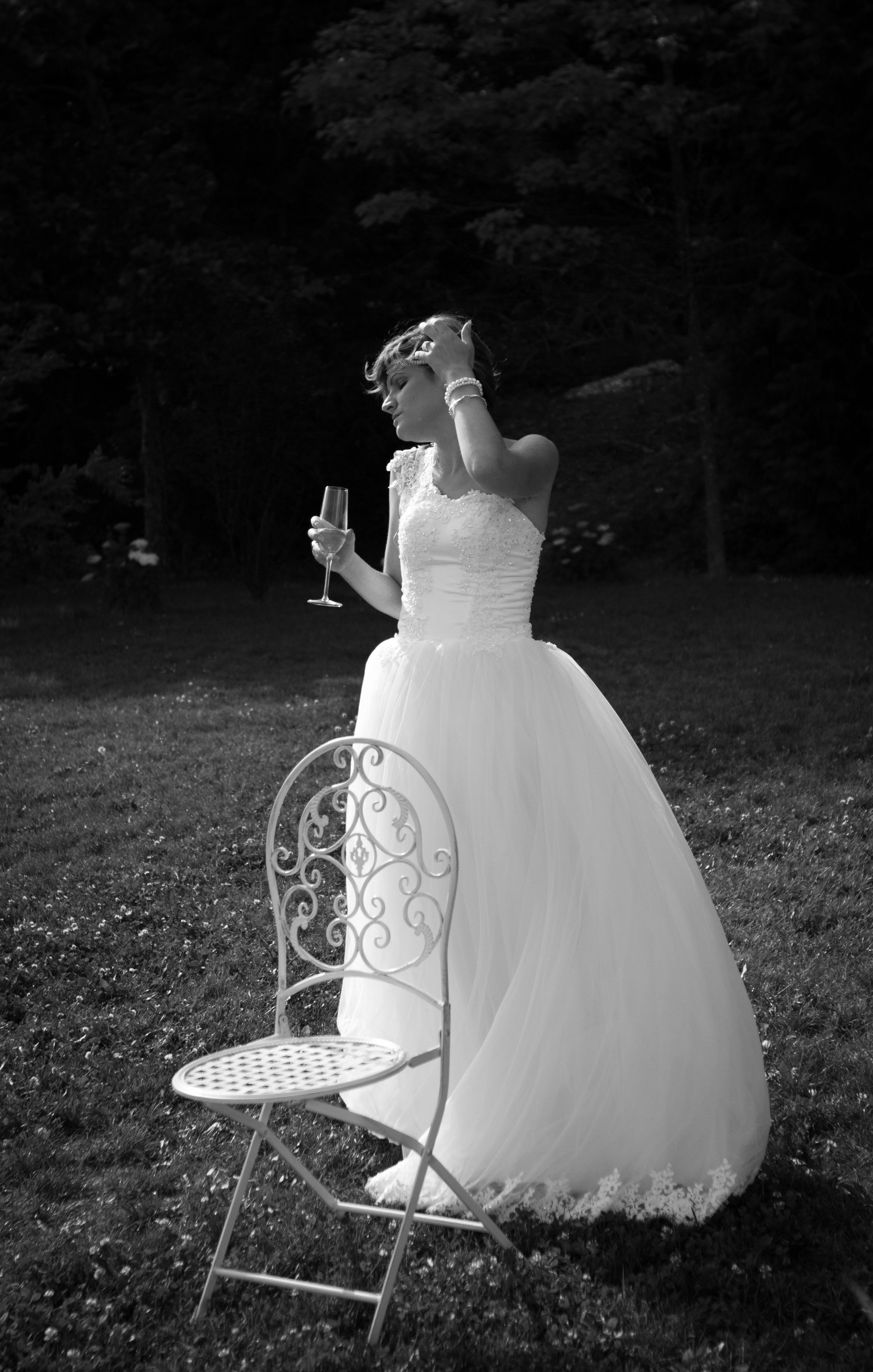 Eine Frau in einem weißen Brautkleid hält ein Champagnerglas in der Hand und berührt ihr Haar mit der anderen Hand, im Hintergrund dichtes Laub und Bäume, im Vordergrund ein leeres, verzierter Gartenstuhl.