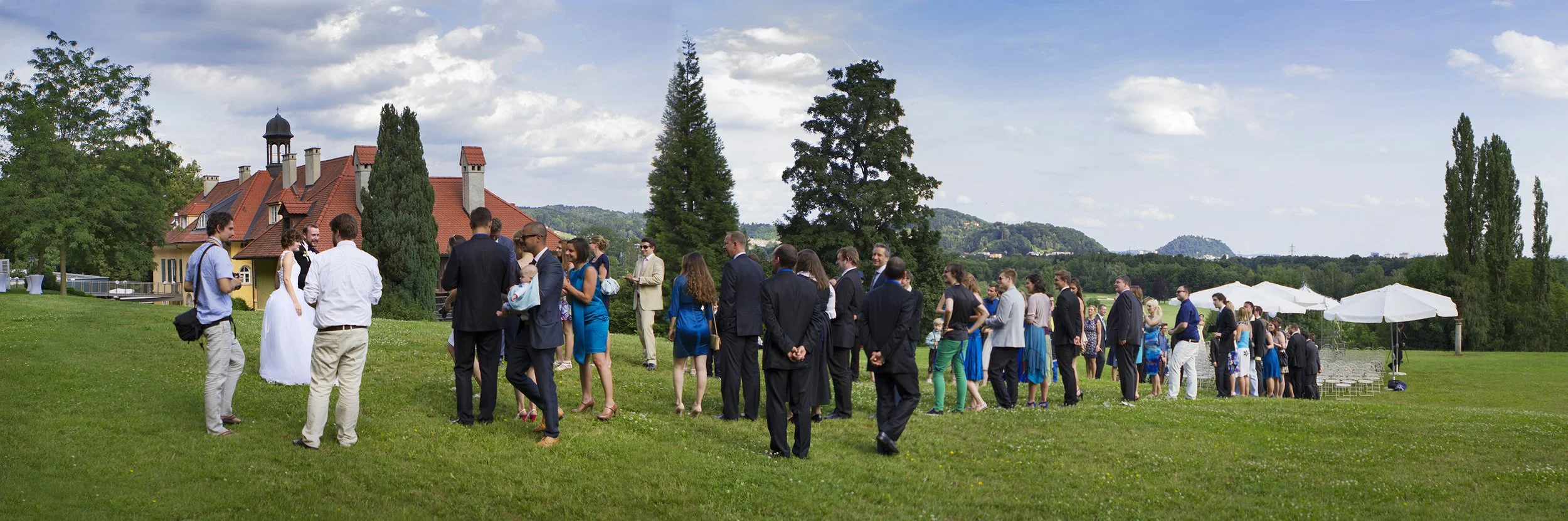 Gruppen von Menschen in formeller Kleidung bei einer outdoor Veranstaltung auf einem grünen Rasen, mit Bäumen und einem historischen Gebäude im Hintergrund, Sonnenschein und Wolken am Himmel.