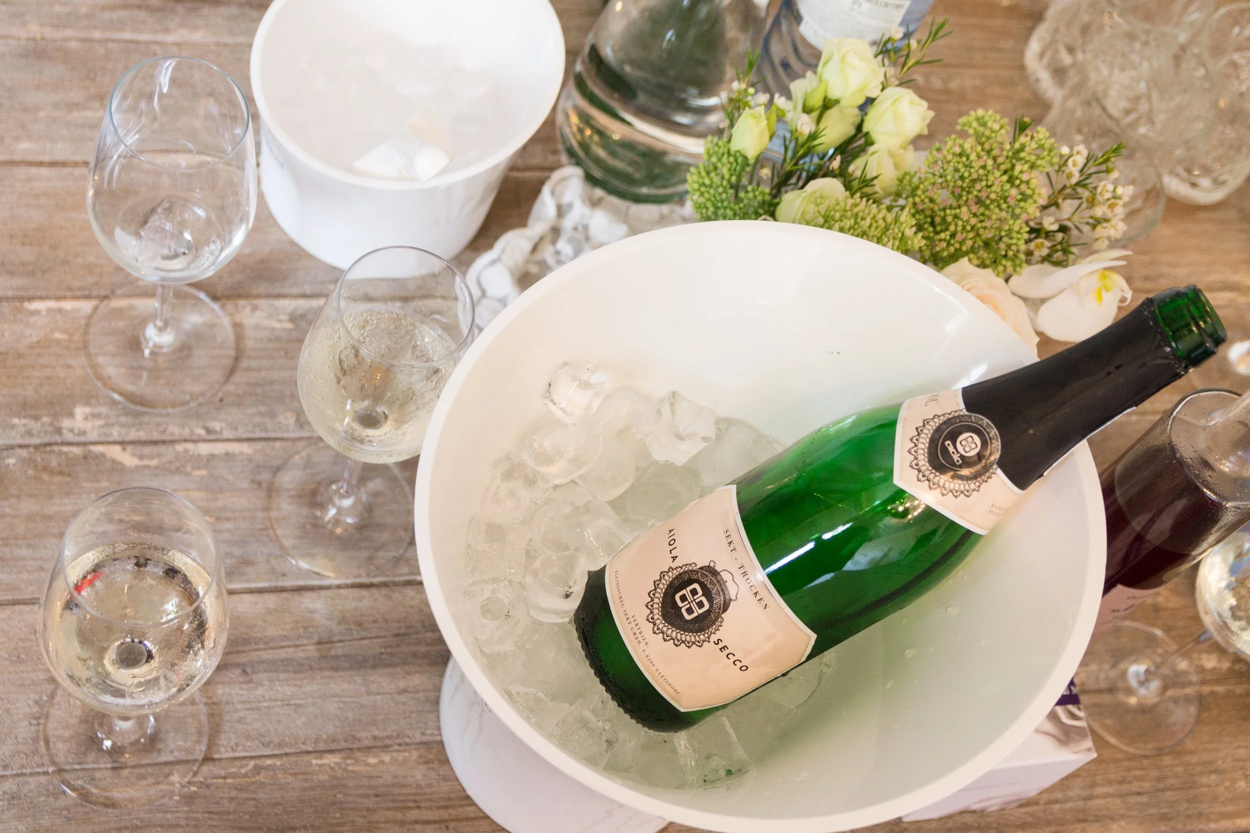 Eine Flasche Sekt in einem Eimer mit Eis, umgeben von mehreren leeren Weingläsern, auf einem Holztisch mit einer Blumen- und Wasserflasche im Hintergrund.