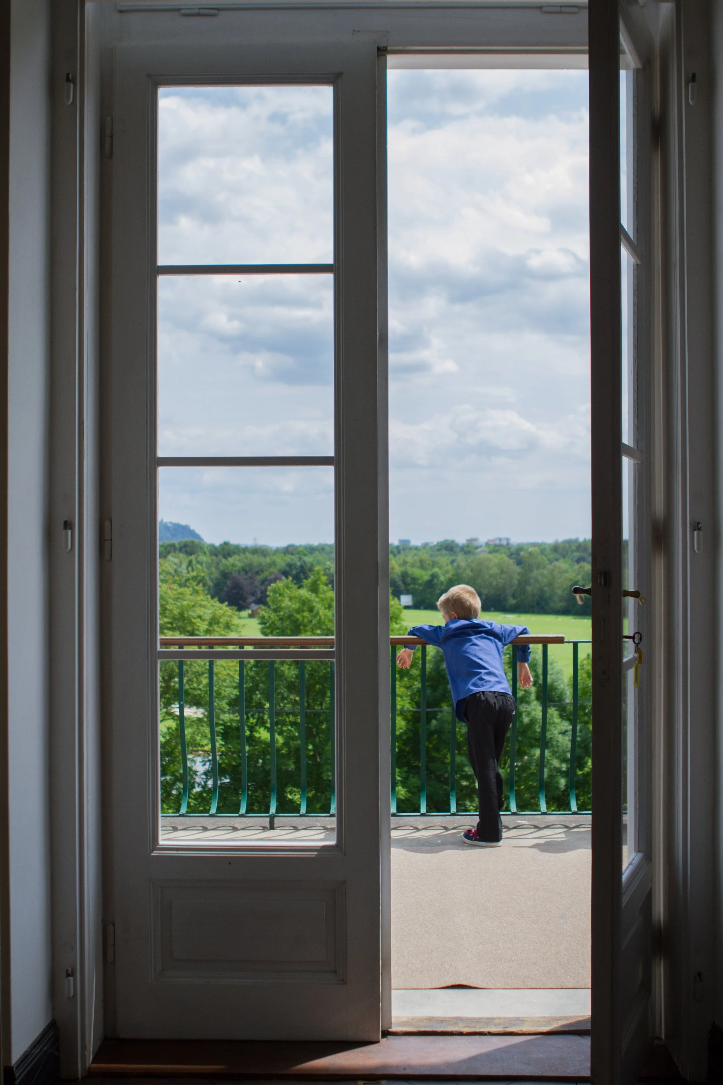 Ein Junge in blauer Jacke und schwarzer Hose steht auf einem Balkon und schaut nach draußen durch eine offene Tür, die zu einem grünen Park mit Bäumen führt.