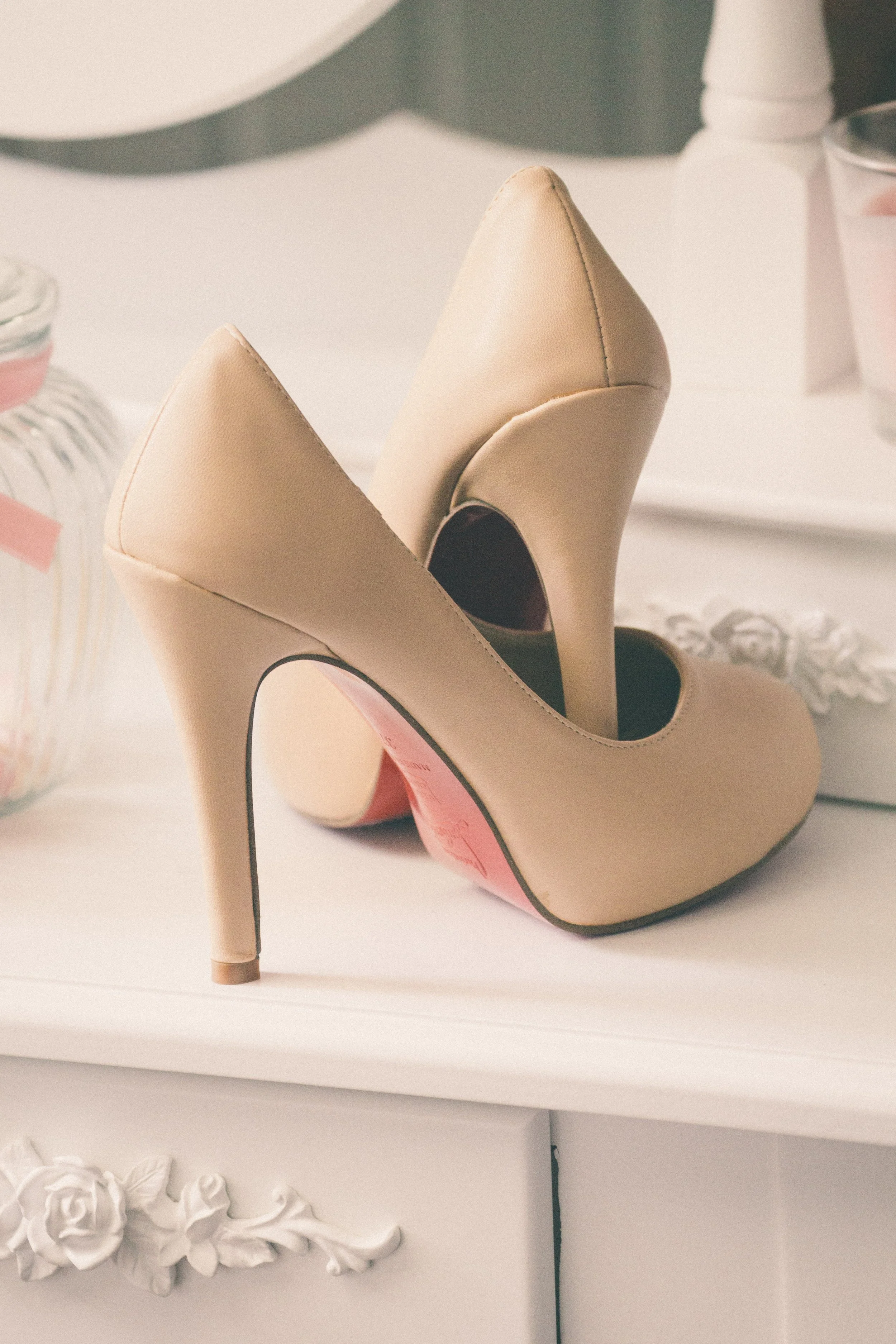 Ein Paar beige High Heels auf einem weißen Regal, umgeben von dekorativen Gegenständen und Deko im Hintergrund.