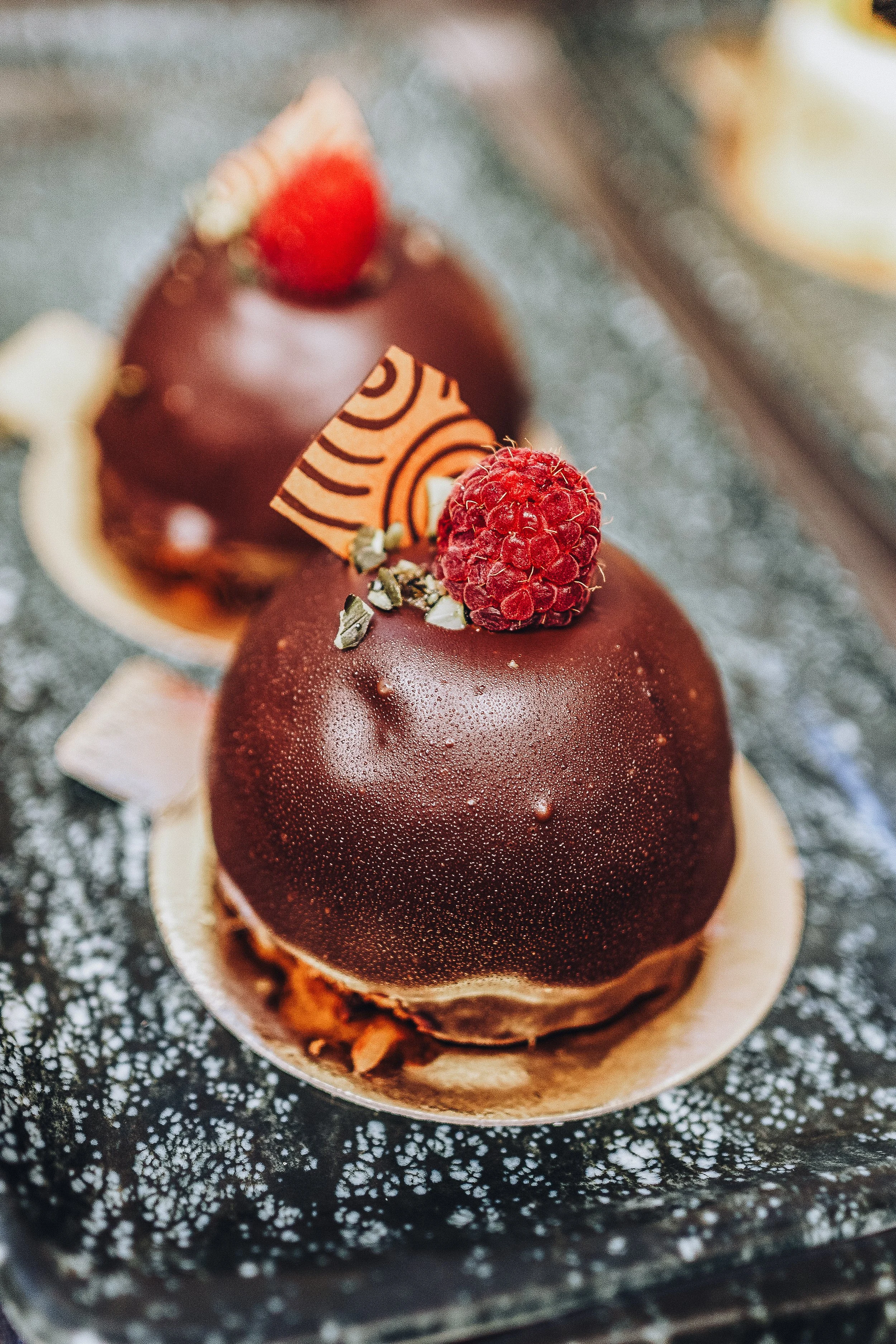Nahaufnahme von zwei eleganten, runden, schokoladigen Desserts mit einer Glasur, verziert mit Himbeeren, Schokolade, weißer Schokolade und kleinen grünen Dekorationen, auf einem dunklen, ornamentalen Hintergrund.