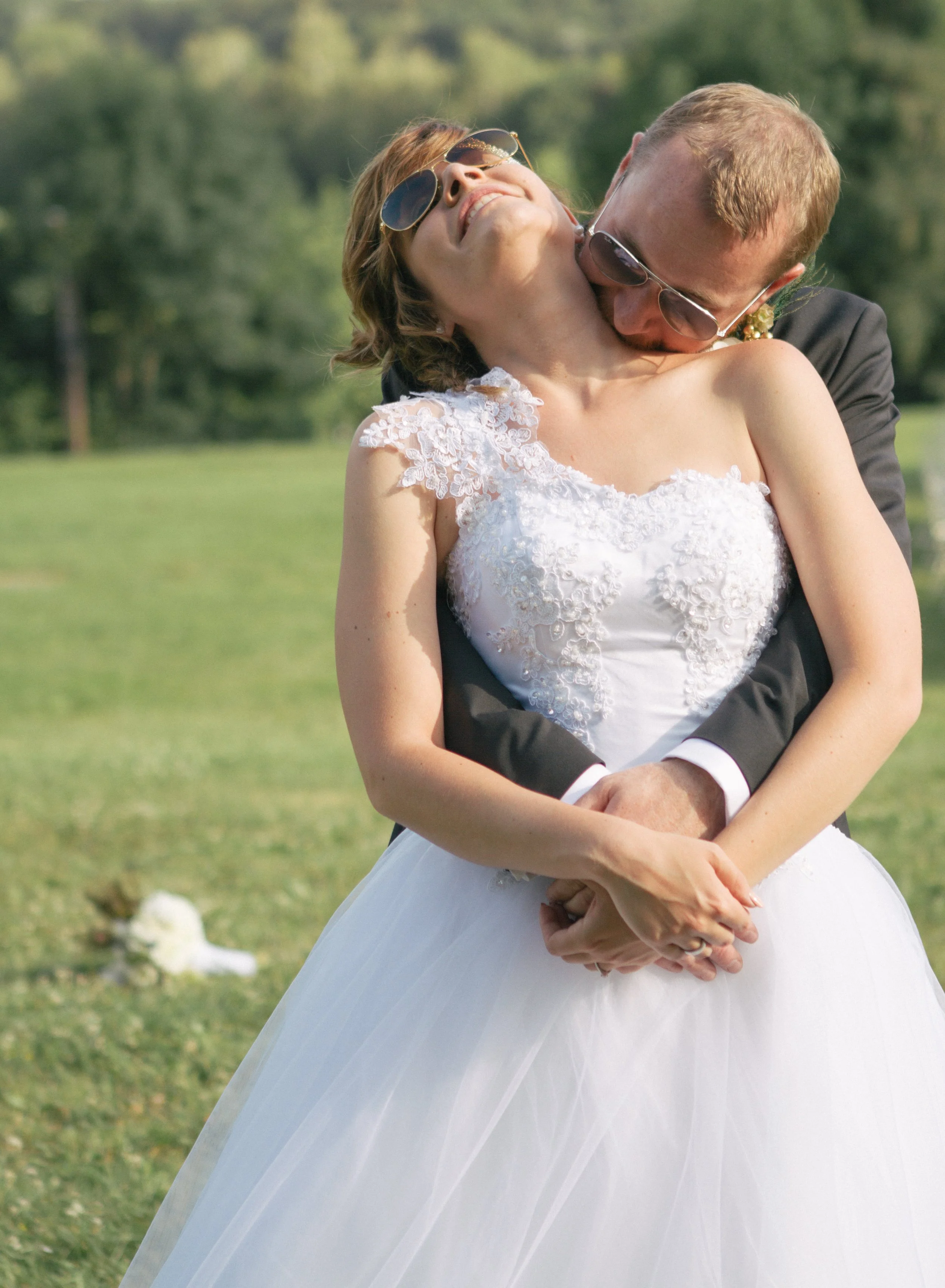 Braut und Bräutigam umarmen sich auf einer grünen Wiese, beide tragen Sonnenbrillen, die Braut trägt ein weißes Hochzeitskleid mit Spitzenärmeln, der Bräutigam trägt einen schwarzen Anzug.