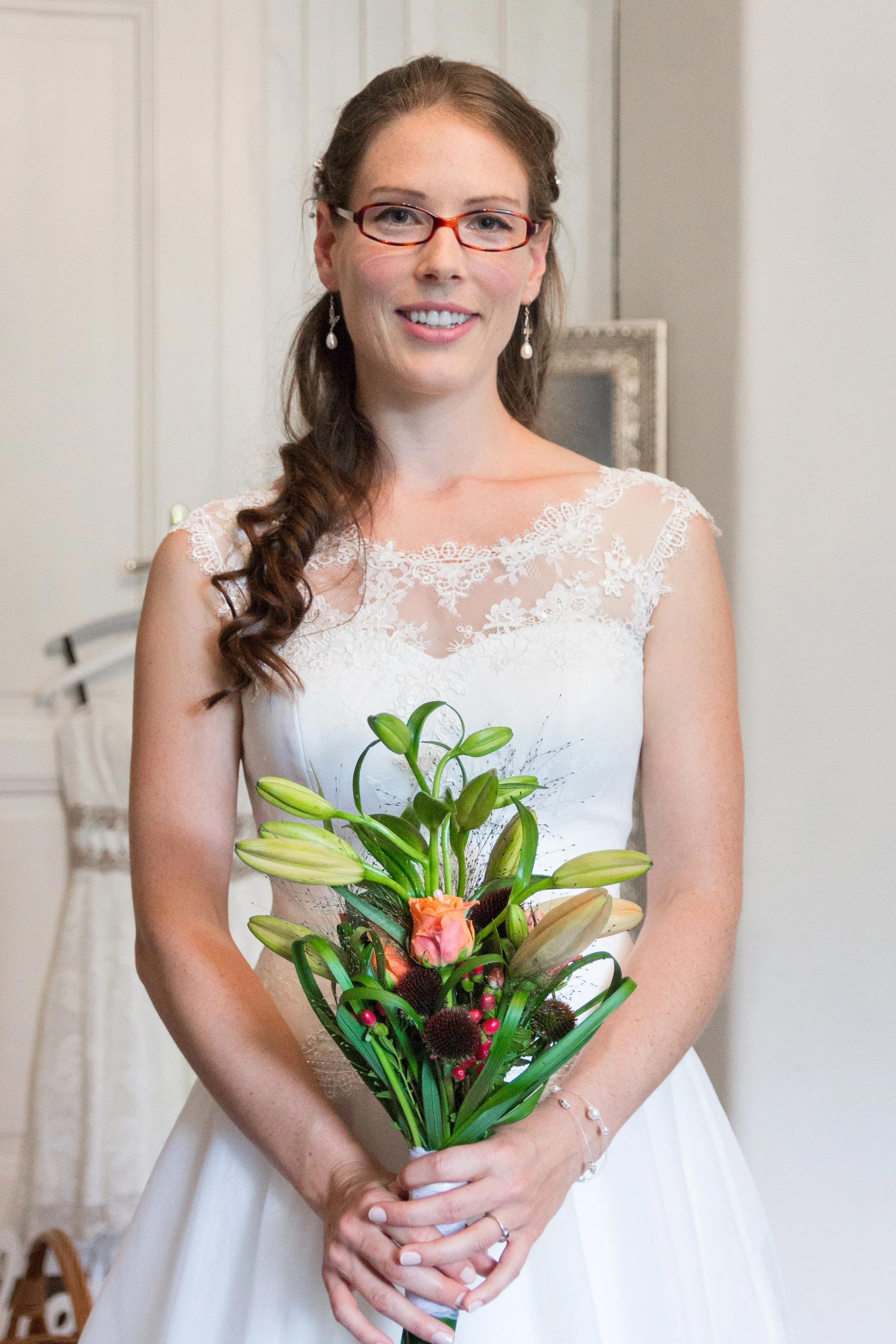 Eine Frau in einem weißen Brautkleid hält einen Blumenstrauß mit Lilien, rosa und dunklen Blumen, vor einem neutralen Hintergrund.