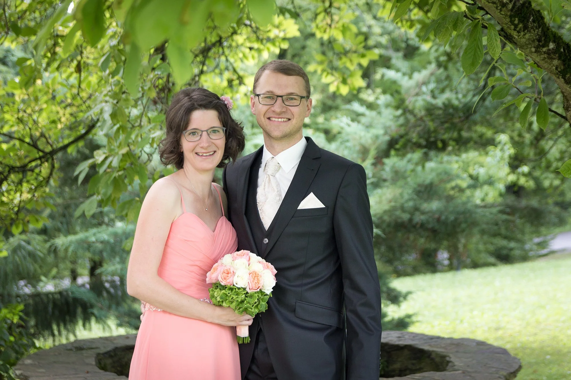 Ein glückliches Brautpaar steht im Grünen. Die Frau trägt ein rosa Brautkleid und hält einen Blumenstrauß. Der Mann trägt einen schwarzen Anzug mit einer weißen Krawatte. Beide lächeln in die Kamera.