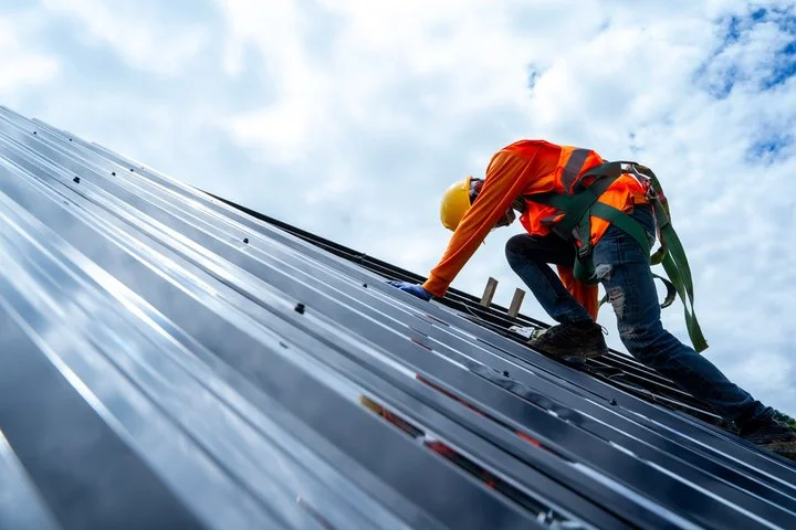 A construction worker wearing an orange safety vest, helmet, and harness, climbing a metal roof against a partly cloudy sky.