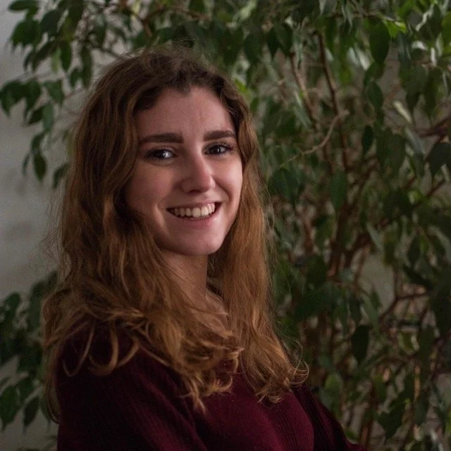 Jeune femme avec des cheveux marron ondulés, souriant, portant un pull bordeaux. En arrière-plan, une plante verte avec des feuilles.